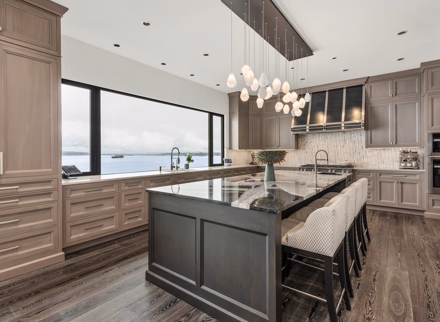 Modern kitchen with large dark island, pendant lights, gray cabinets, and a window overlooking water.