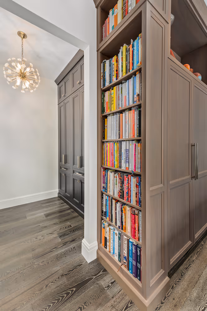 Tall built-in wooden bookshelf filled with books next to a kitchen area with cabinets and chandelier.