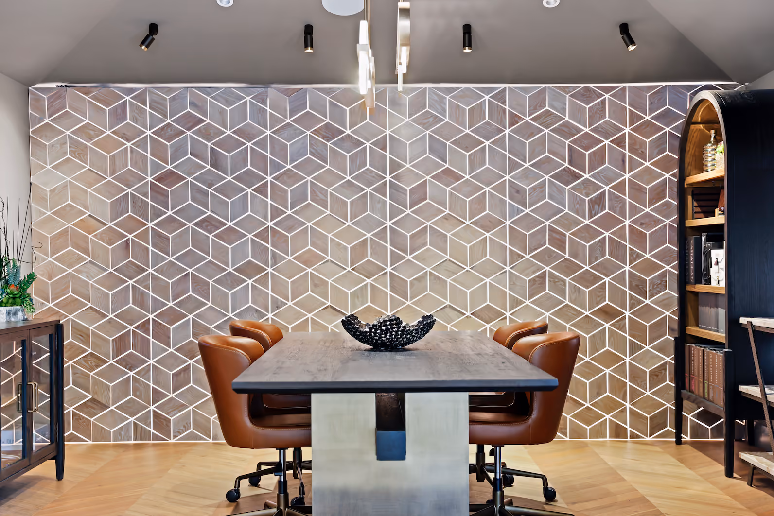 Modern dining room with a rectangular wooden table surrounded by six brown leather chairs on wheels, a decorative bowl centerpiece, geometric patterned accent wall, wooden floor, and shelves with books and décor on the sides.
