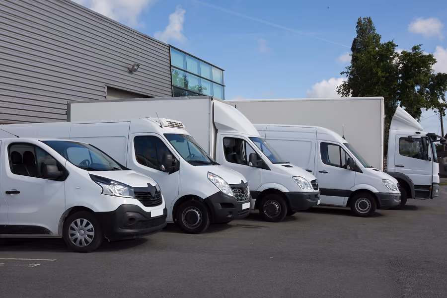 Row of five white commercial vans and trucks parked outside a building under a blue sky.