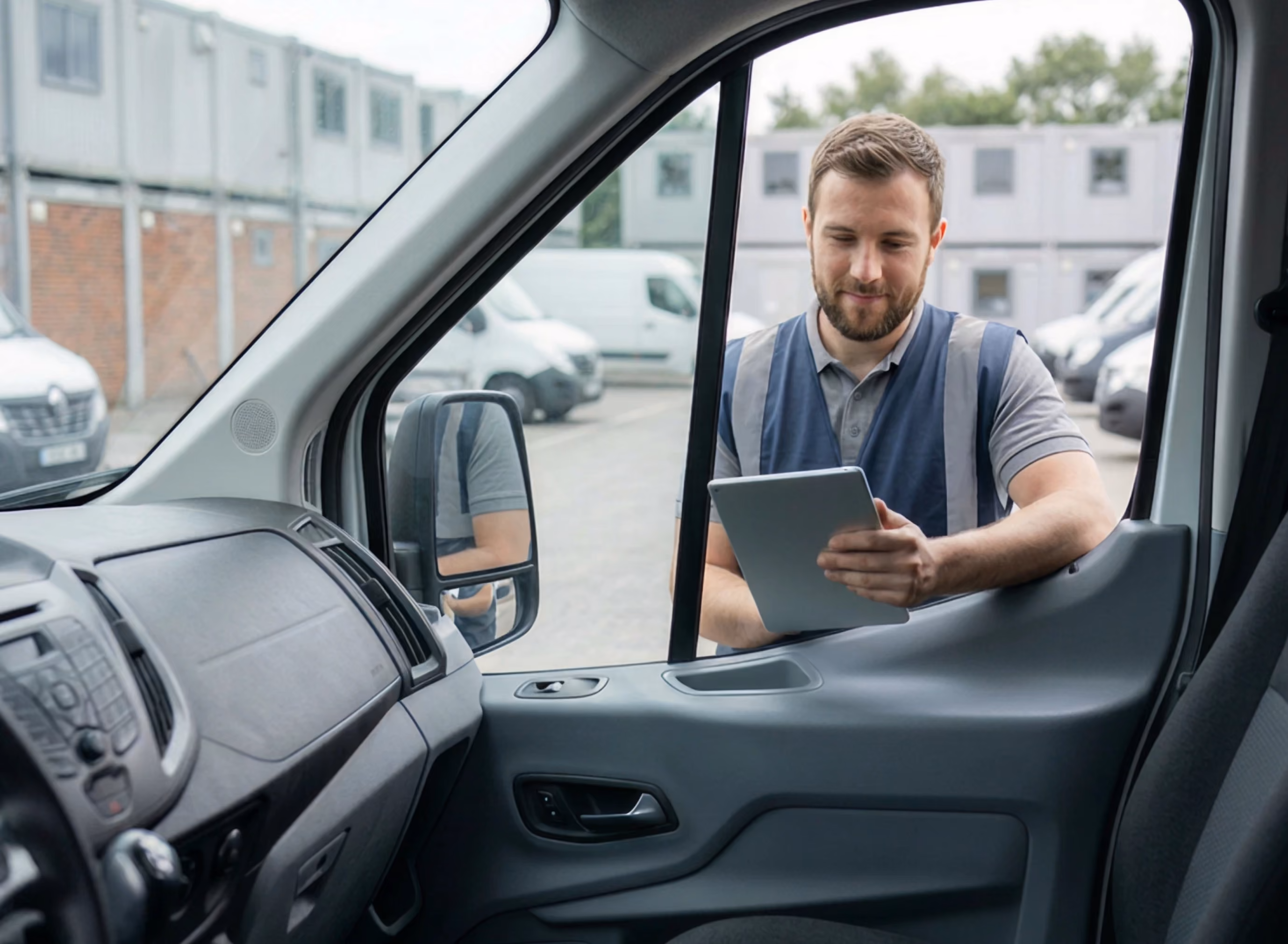 Man in safety vest standing outside a van, looking at a tablet near the driver's window.