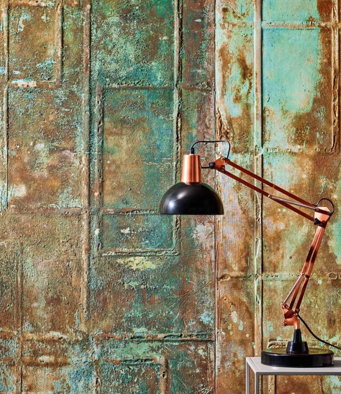 Copper and black adjustable desk lamp on a small table against a textured rusted metal wall.