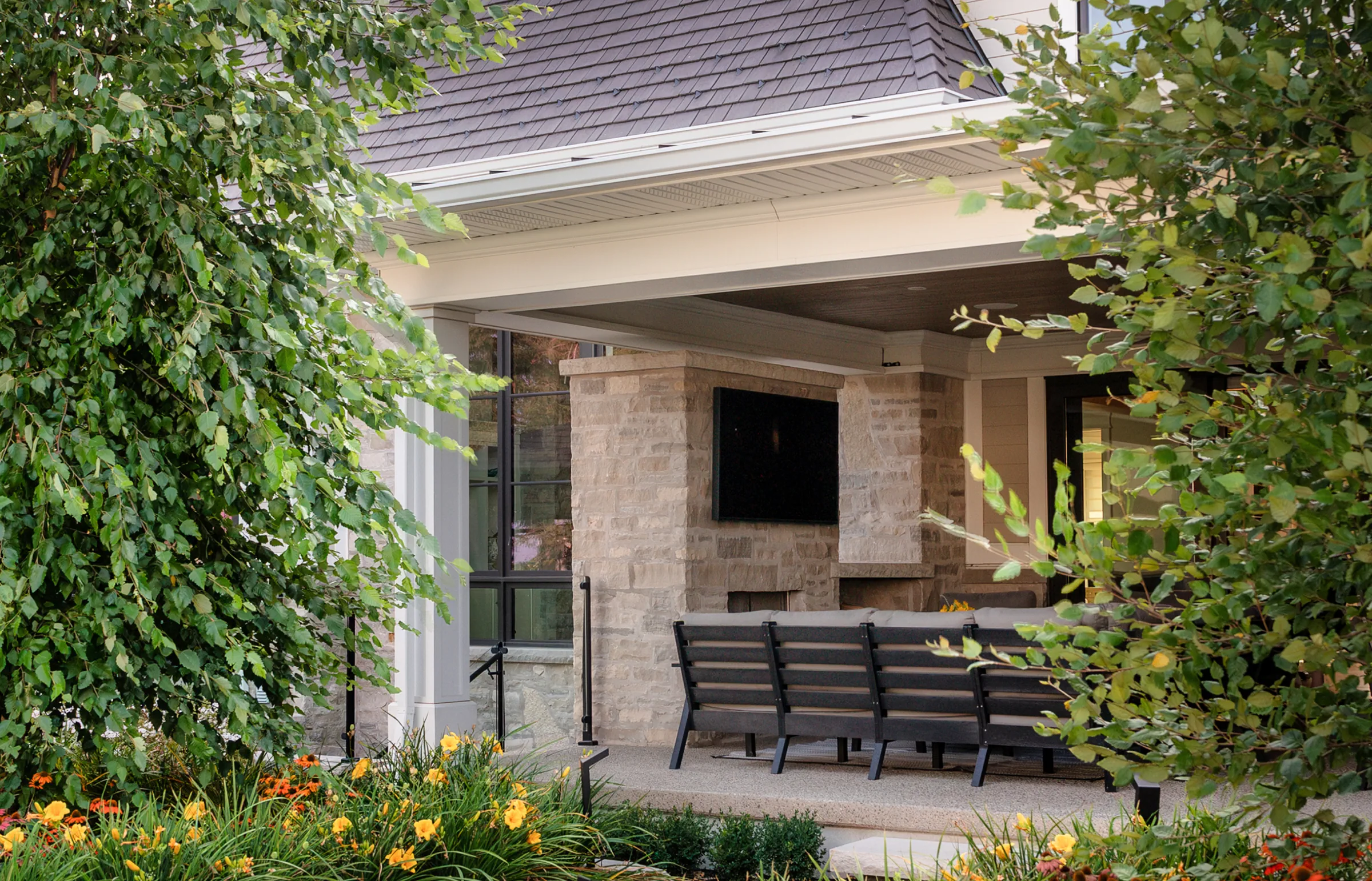 Covered patio beneath a grey shake-style roof, with stone columns and garden planting.