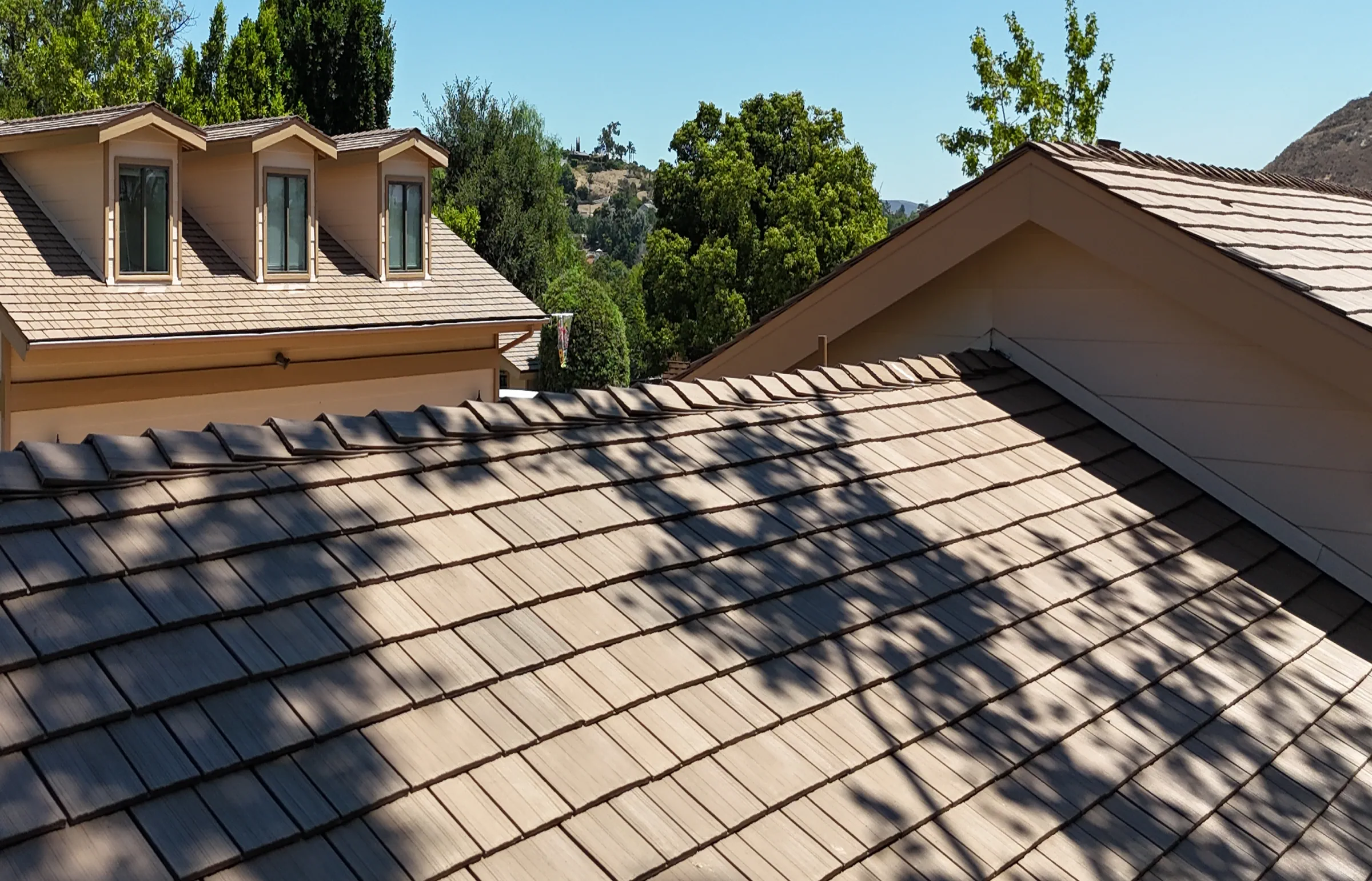 Tan shake-style roofing in staggered rows with tree shadows and three dormers in the background.