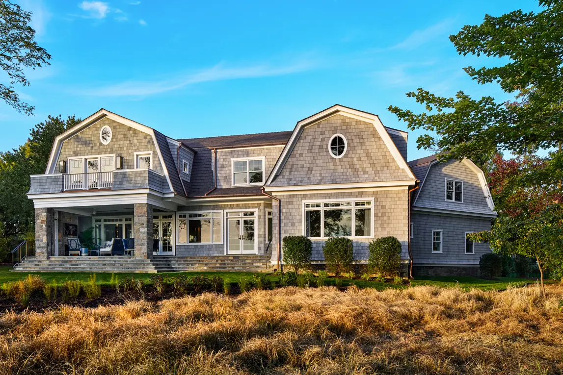Large gray shingle-style home with multiple gambrel roofs in a gray Enviroshake composite shake, oval windows, a stone-columned porch, and coastal grasses in the foreground.