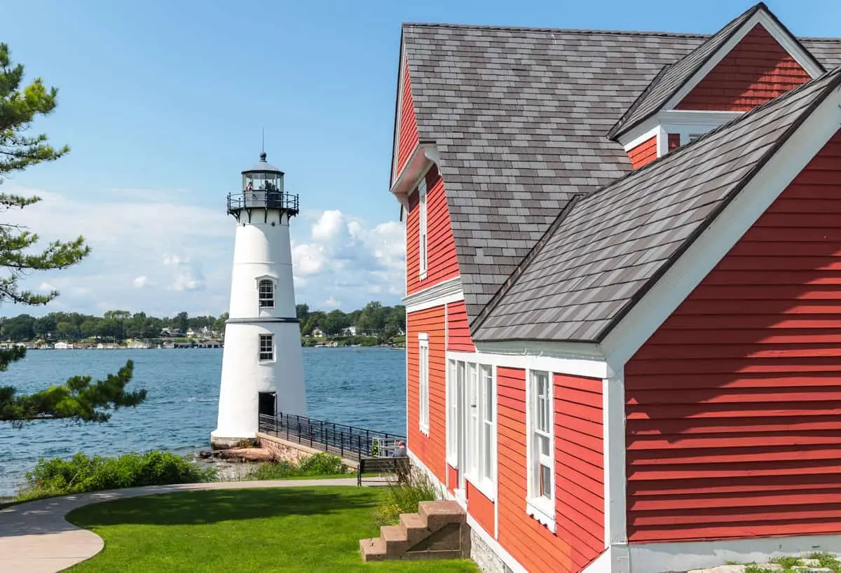 Historic red keeper's house with an Enviroshake composite roof in a mixed-gray shake style, overlooking a white lighthouse and waterfront on a clear day.