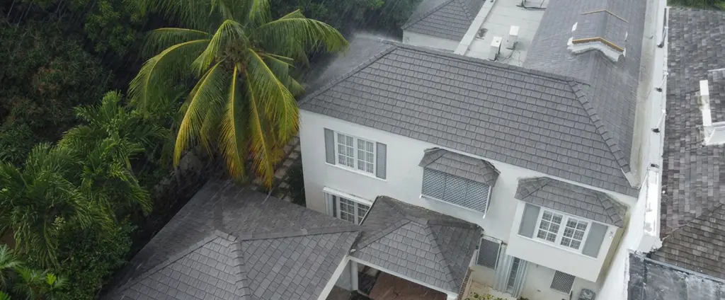 Aerial view of a two-story home during rainfall, featuring an Enviroshake composite roof in a weathered gray shake style, surrounded by tropical palm trees.