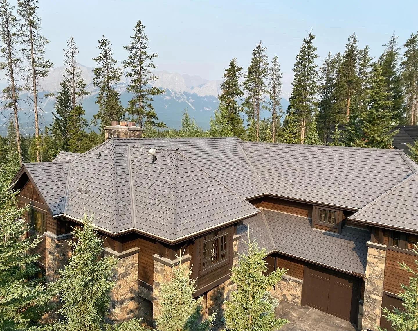 Aerial view of a large mountain home with a gray Enviroshake composite roof, stone and timber exterior, surrounded by pine trees with a mountain range in the background.