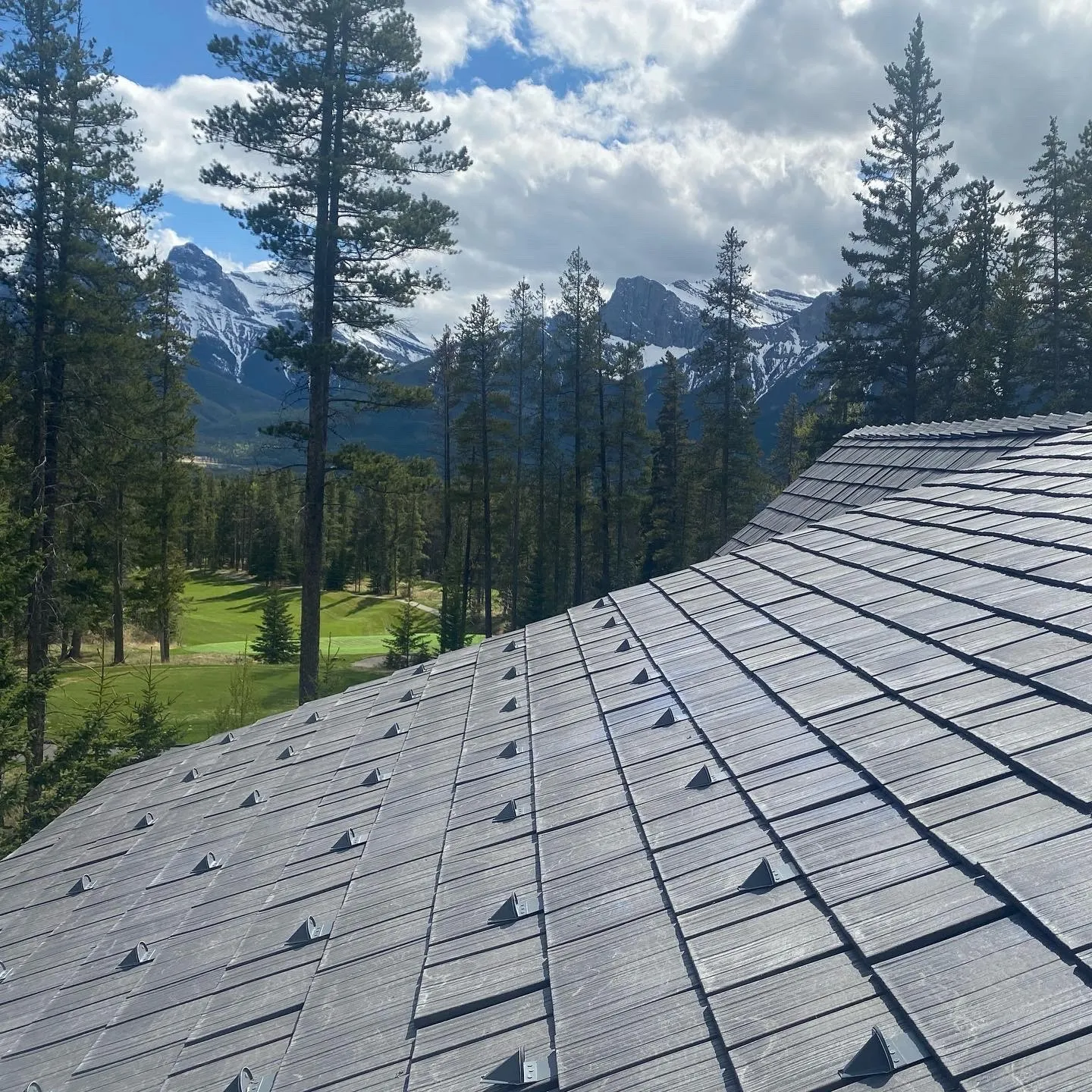 Close-up view across a gray Enviroshake composite roof with snow guards, overlooking pine trees, a golf course, and snow-capped mountains.