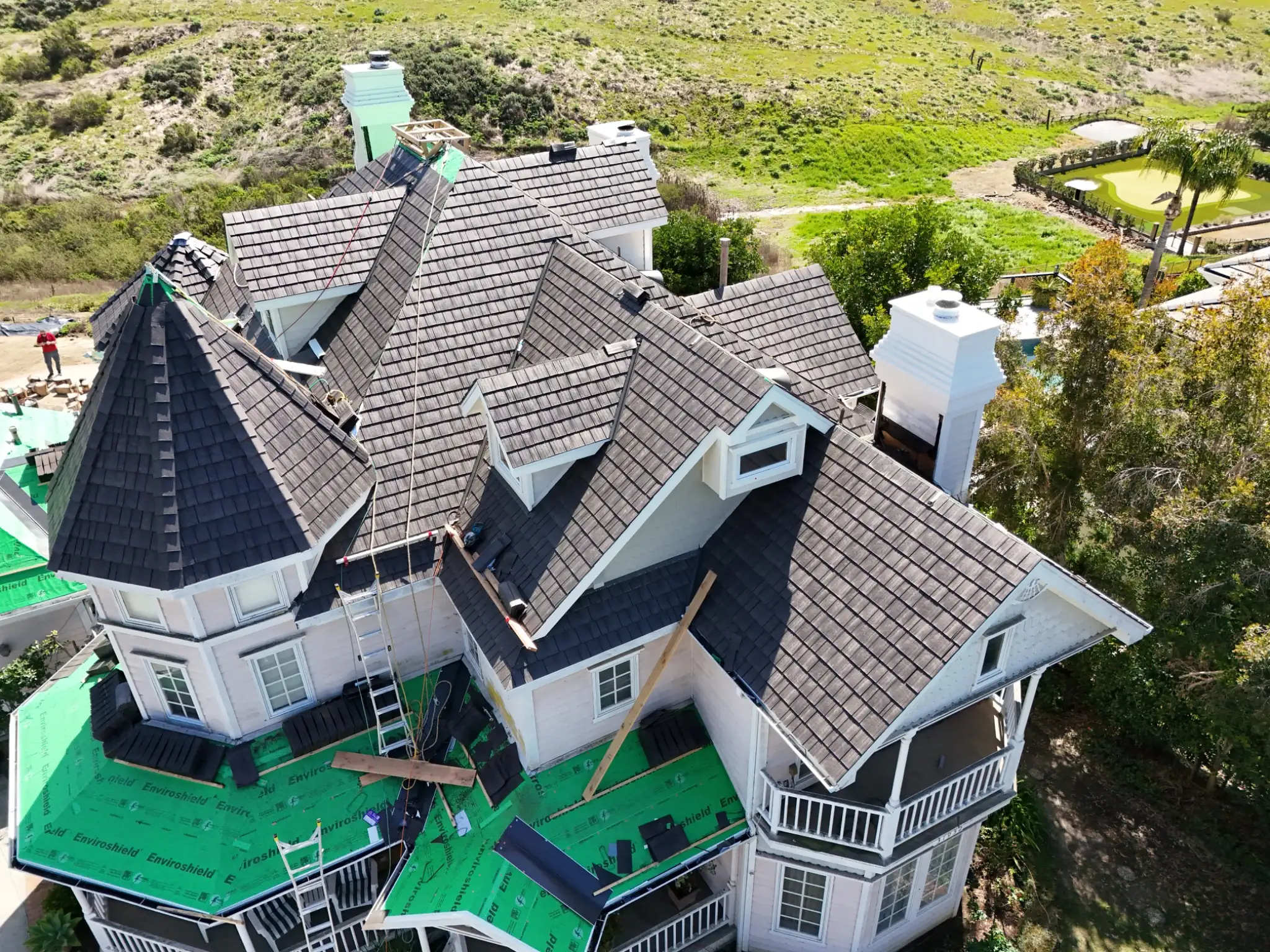 Aerial view of a Victorian-style home mid-installation of a gray Enviroshake composite roof, with green Enviroshield underlayment visible, coastal hillside setting