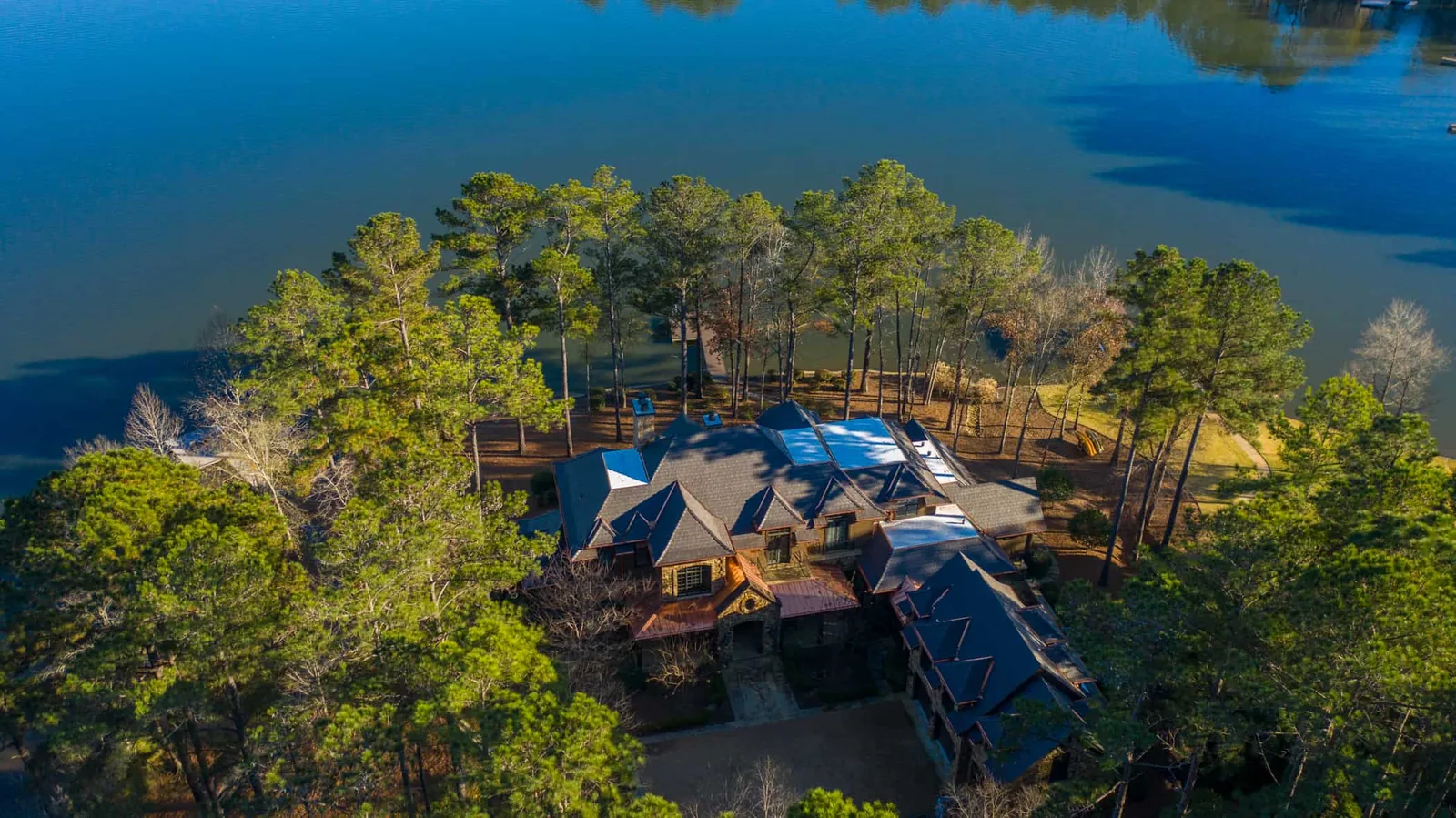 Aerial view of a large stone and timber lakefront estate with a dark gray Enviroshake composite roof, copper roof accents, and skylights, surrounded by pine trees on a peninsula