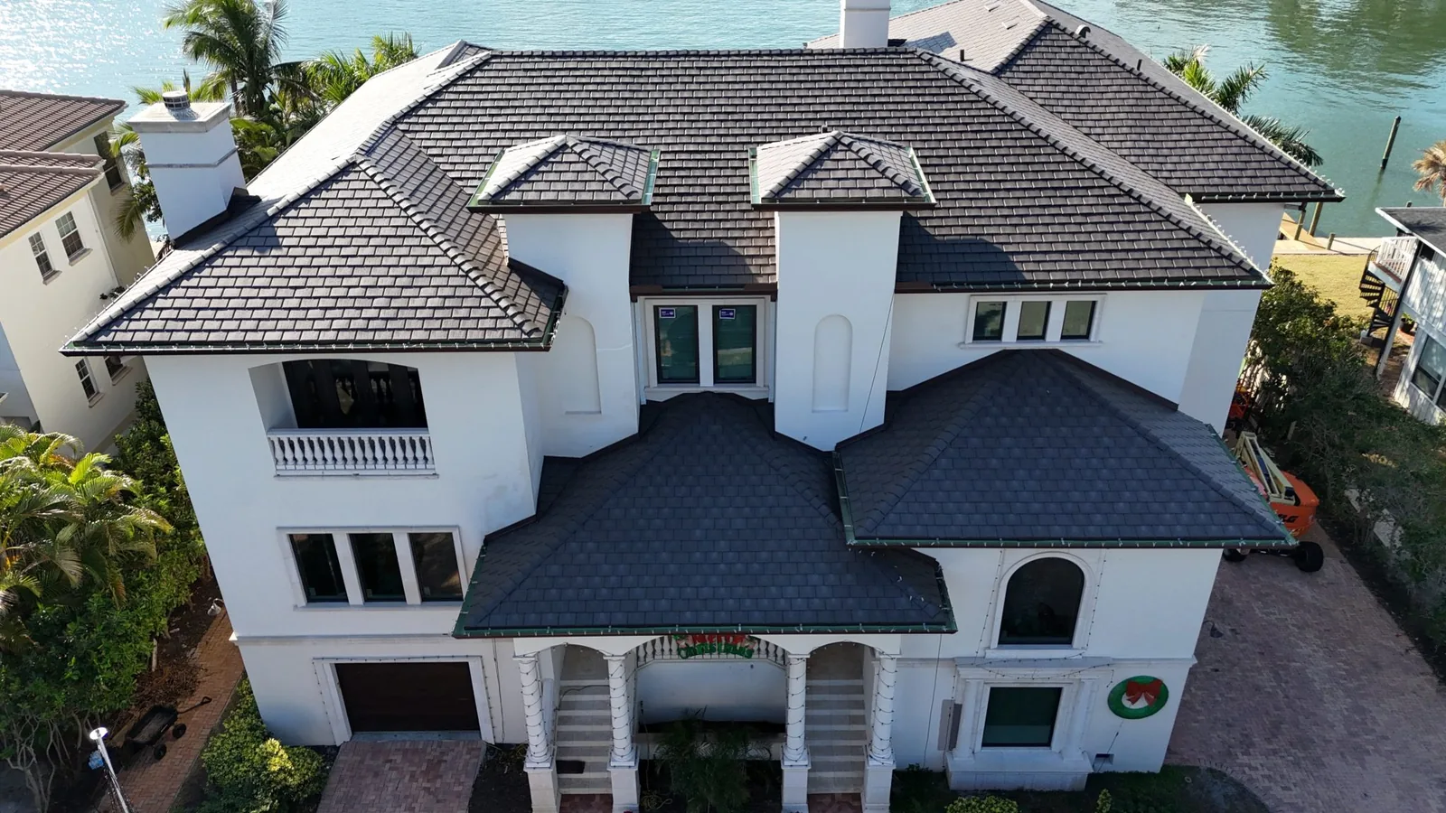 Aerial view of a large white waterfront estate with a black Enviroslate composite slate roof, arched entrance with columns, and holiday lights, along a canal with palm trees.