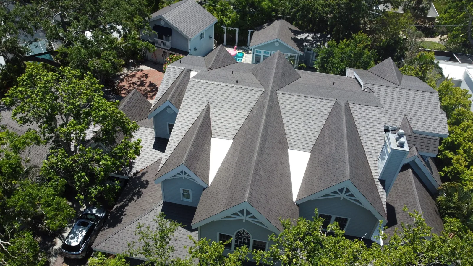 Aerial view of a sprawling coastal home with a light gray Enviroshake composite roof, multiple gables and dormers, a chimney, and a backyard pool, surrounded by dense tree cover