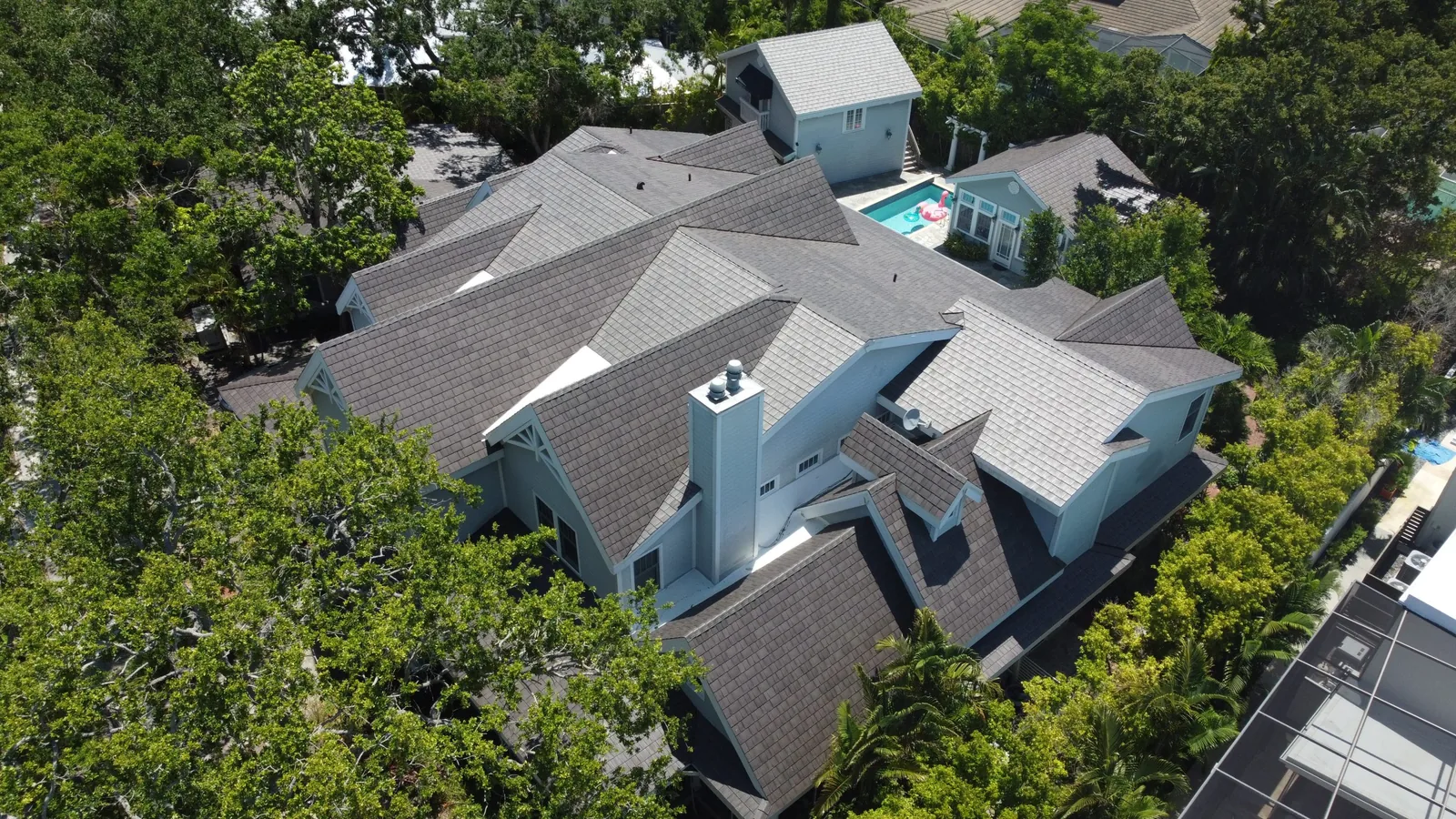 Aerial view of a sprawling coastal home with a light gray Enviroshake composite roof, multiple gables and dormers, a chimney, and a backyard pool, surrounded by dense tree cover