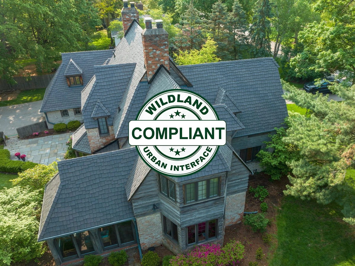 Aerial view of a large tudor-style home with a gray Enviroshake composite roof, brick chimneys, and stone accents, overlaid with a "Wildland Urban Interface Compliant" badge.