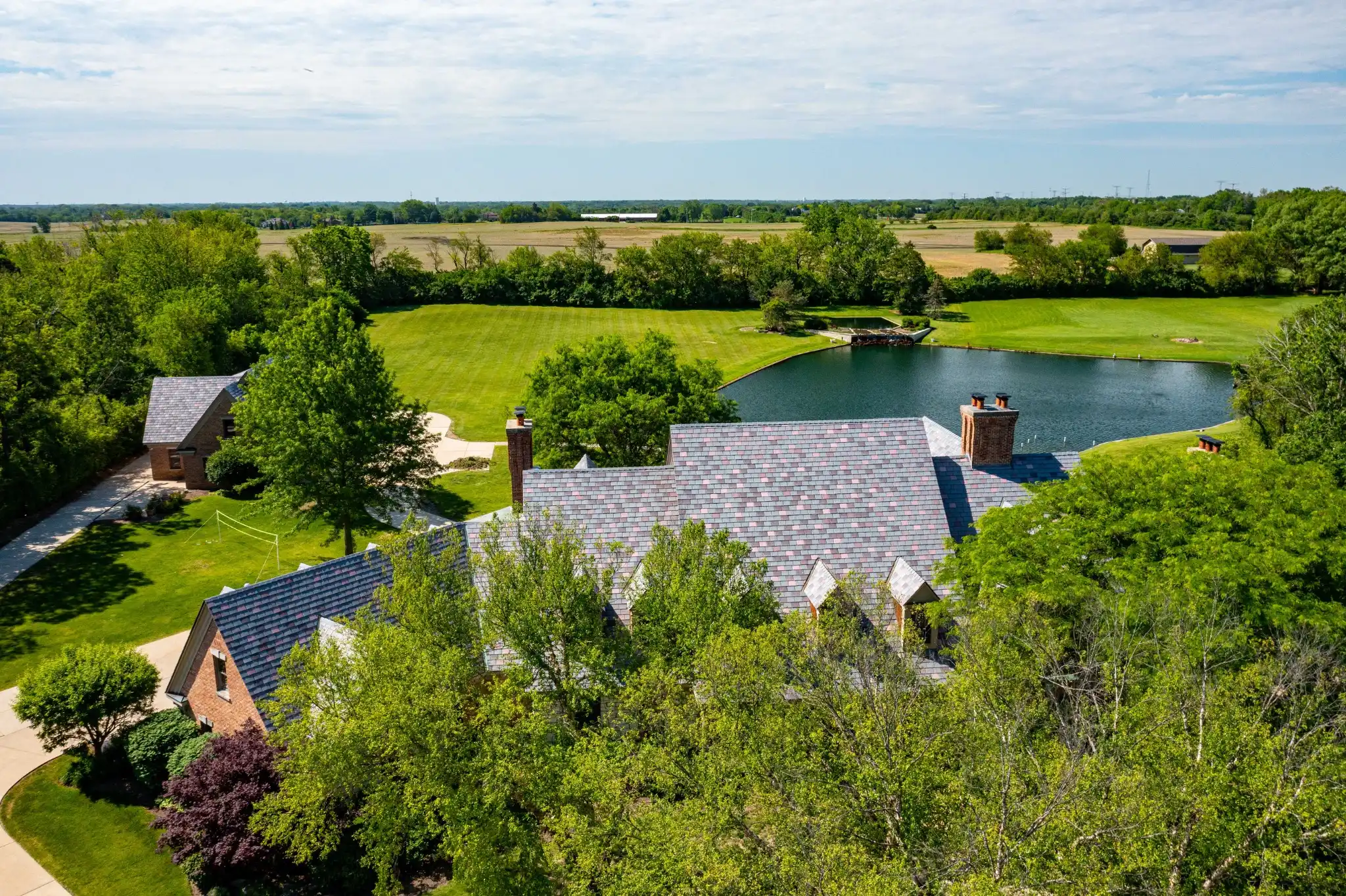 Aerial view of a large brick estate with a multitone Enviroslate composite slate roof in blended purple-gray tones, brick chimneys, and expansive grounds with a private pond and open fields.