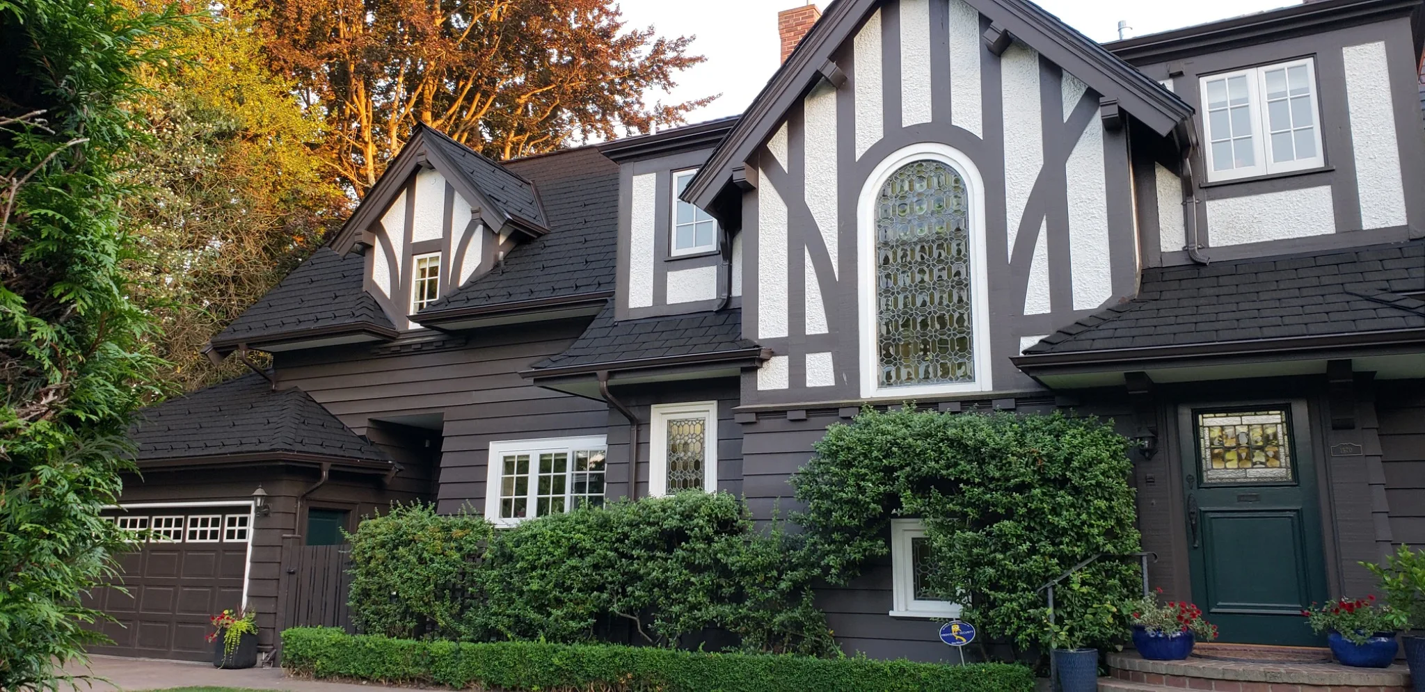 Tudor-style home with a black Enviroslate composite slate roof, dark timber and white stucco exterior, arched leaded-glass windows, and manicured hedges along the front.
