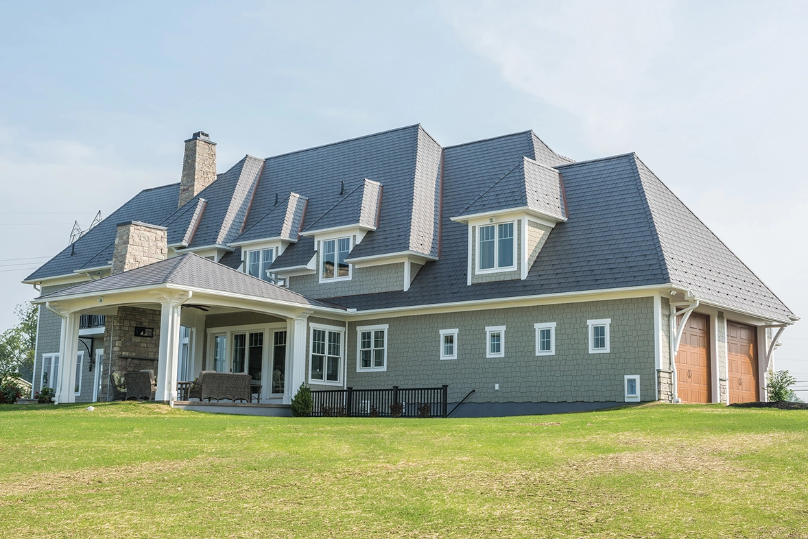 Large home with charcoal gray Enviroslate composite slate roof, multiple dormers, stone chimney, and covered porch, set on an open lawn