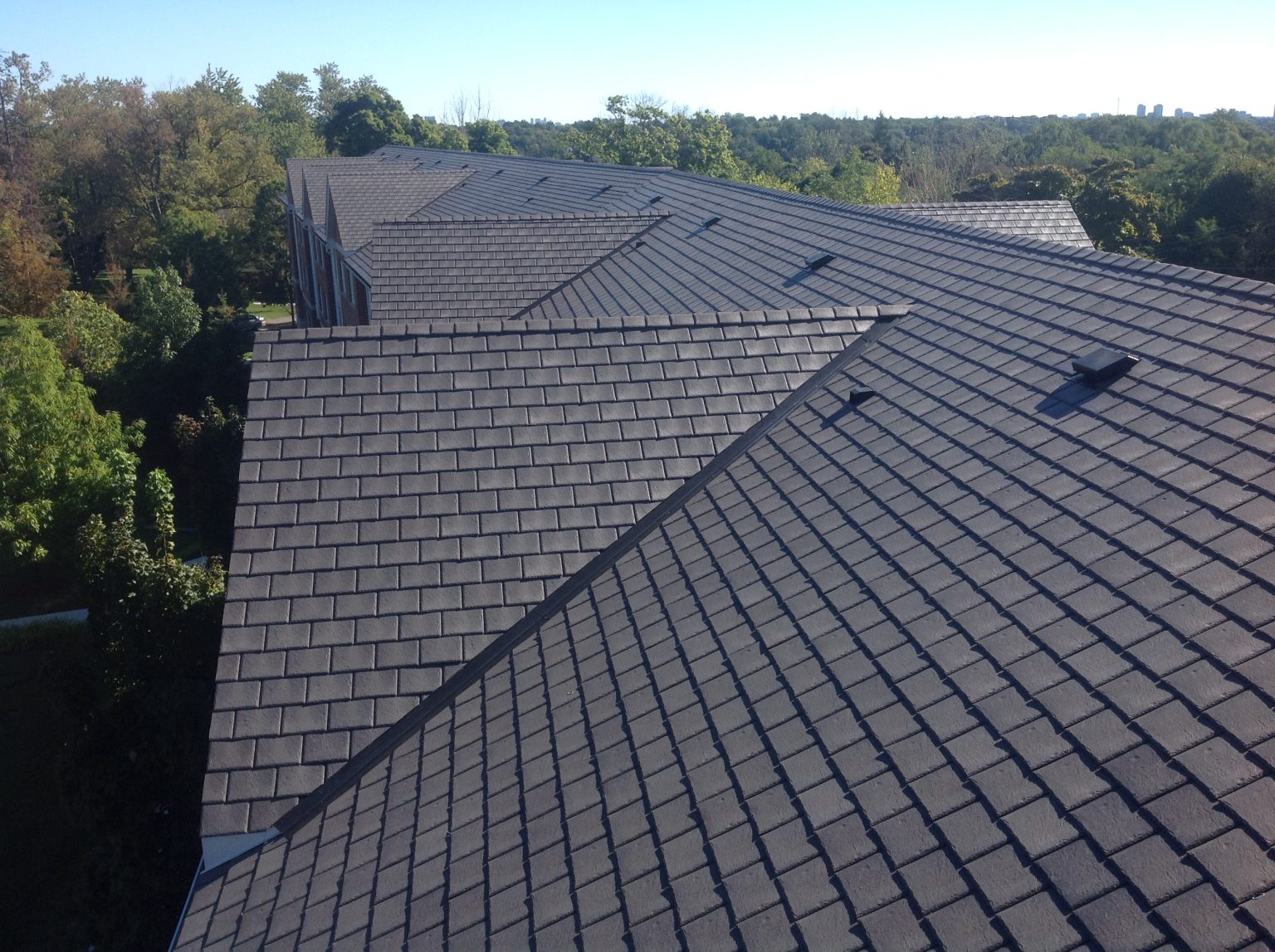  Aerial close-up of a large dark gray Enviroslate composite slate roof spanning a multi-gabled commercial building, with roof vents and a treed skyline in the background.