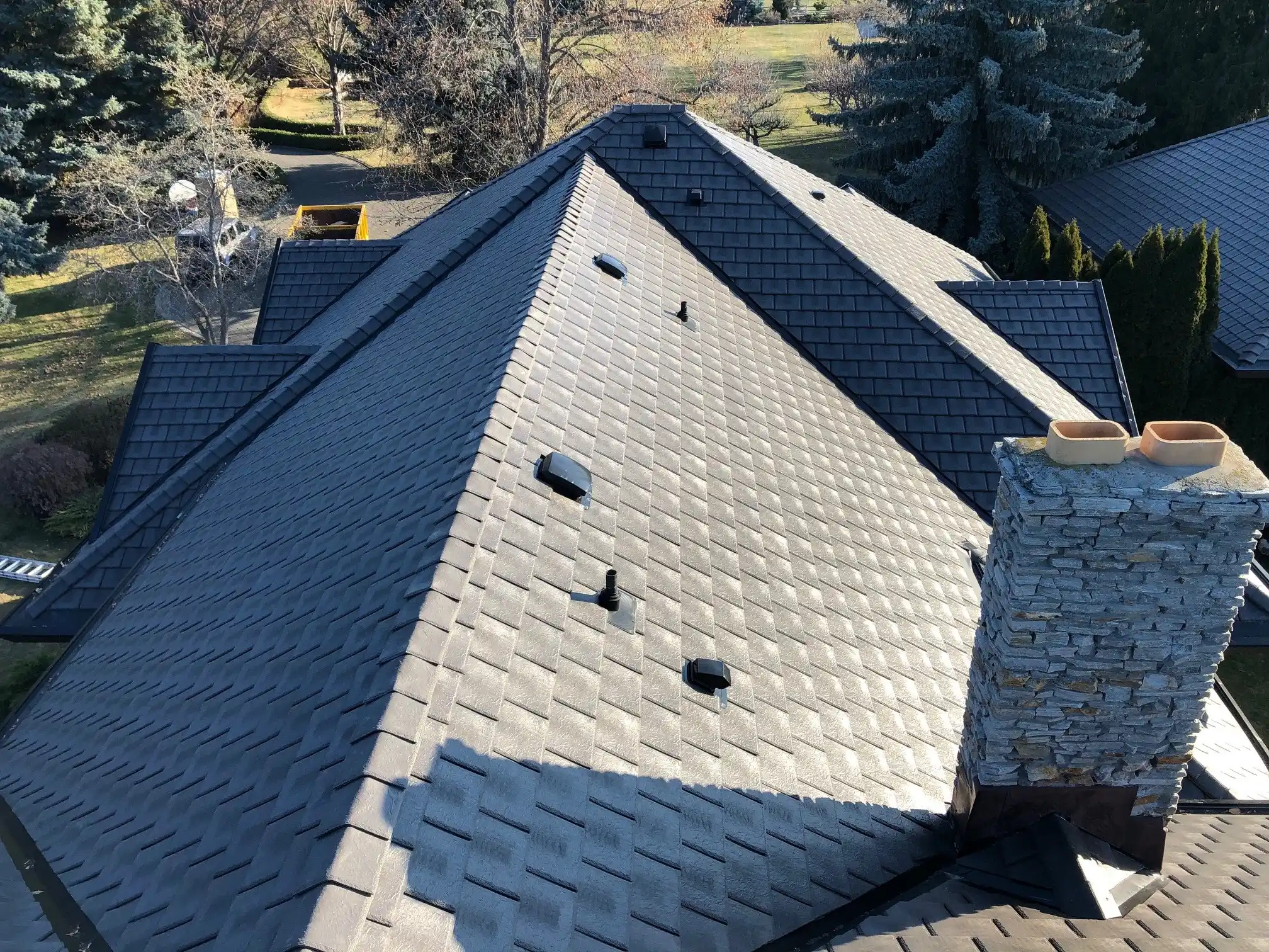 Aerial view of a gray Enviroslate composite slate roof with multiple hips and roof vents, next to a stone chimney with terra cotta chimney pots, overlooking a wooded property.