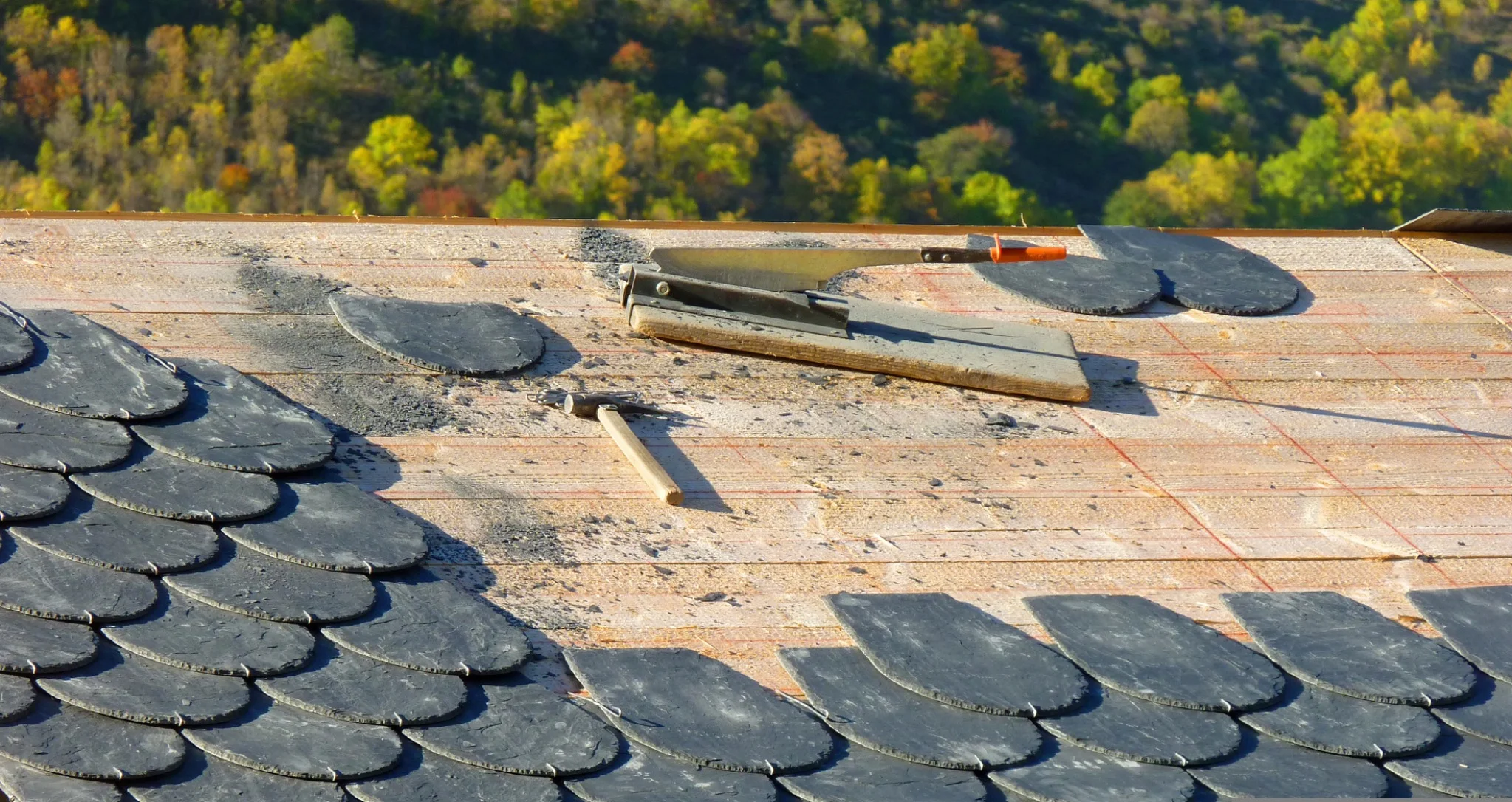 Close-up of a natural slate roof mid-installation, with rounded scalloped tiles laid in a pattern, exposed underlayment, and roofing tools resting on the deck.