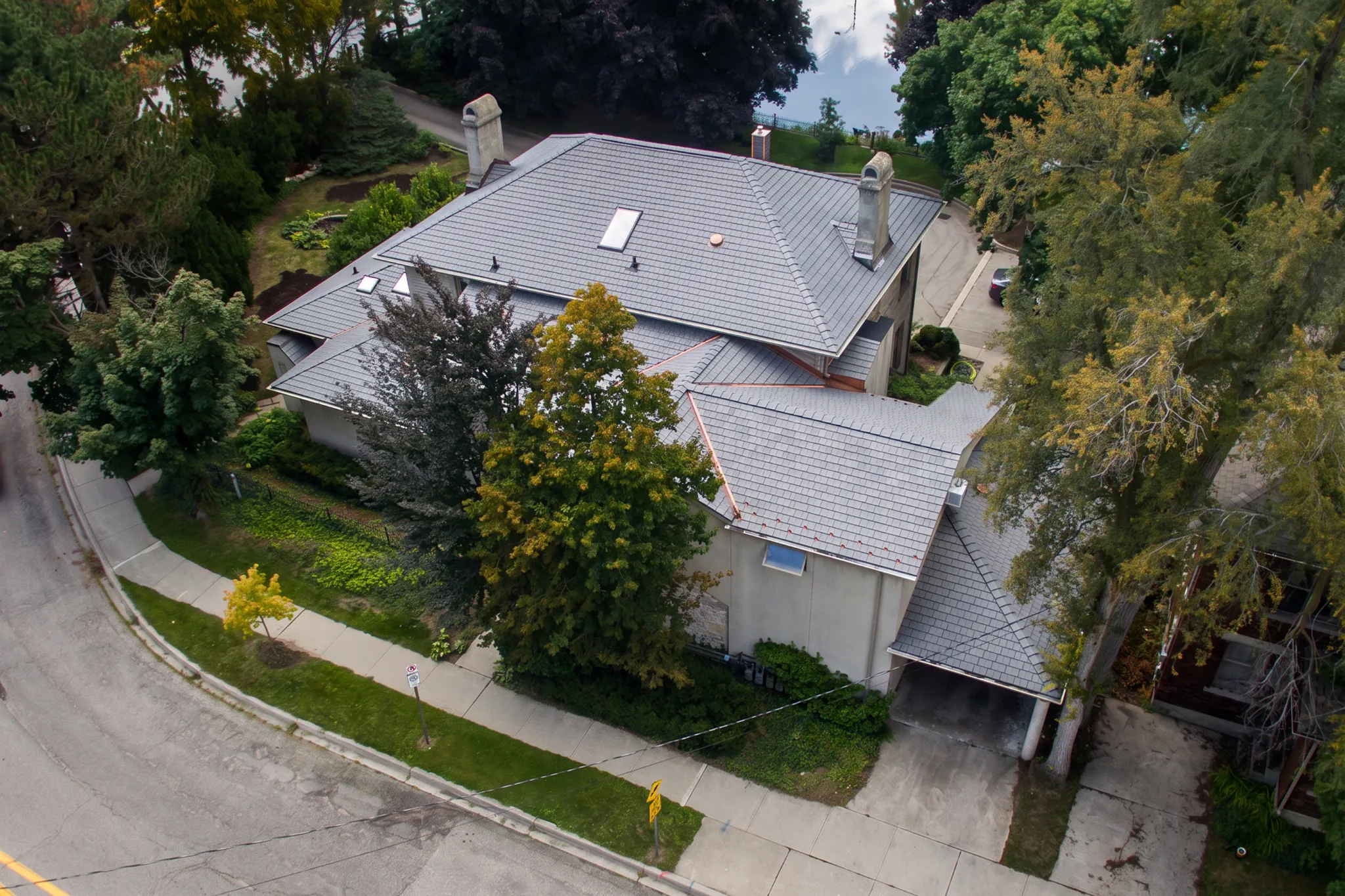 Aerial view of a large gray Enviroshake composite roof on a multi-level home with two chimneys, skylights, and copper flashing, surrounded by mature trees near a residential street.