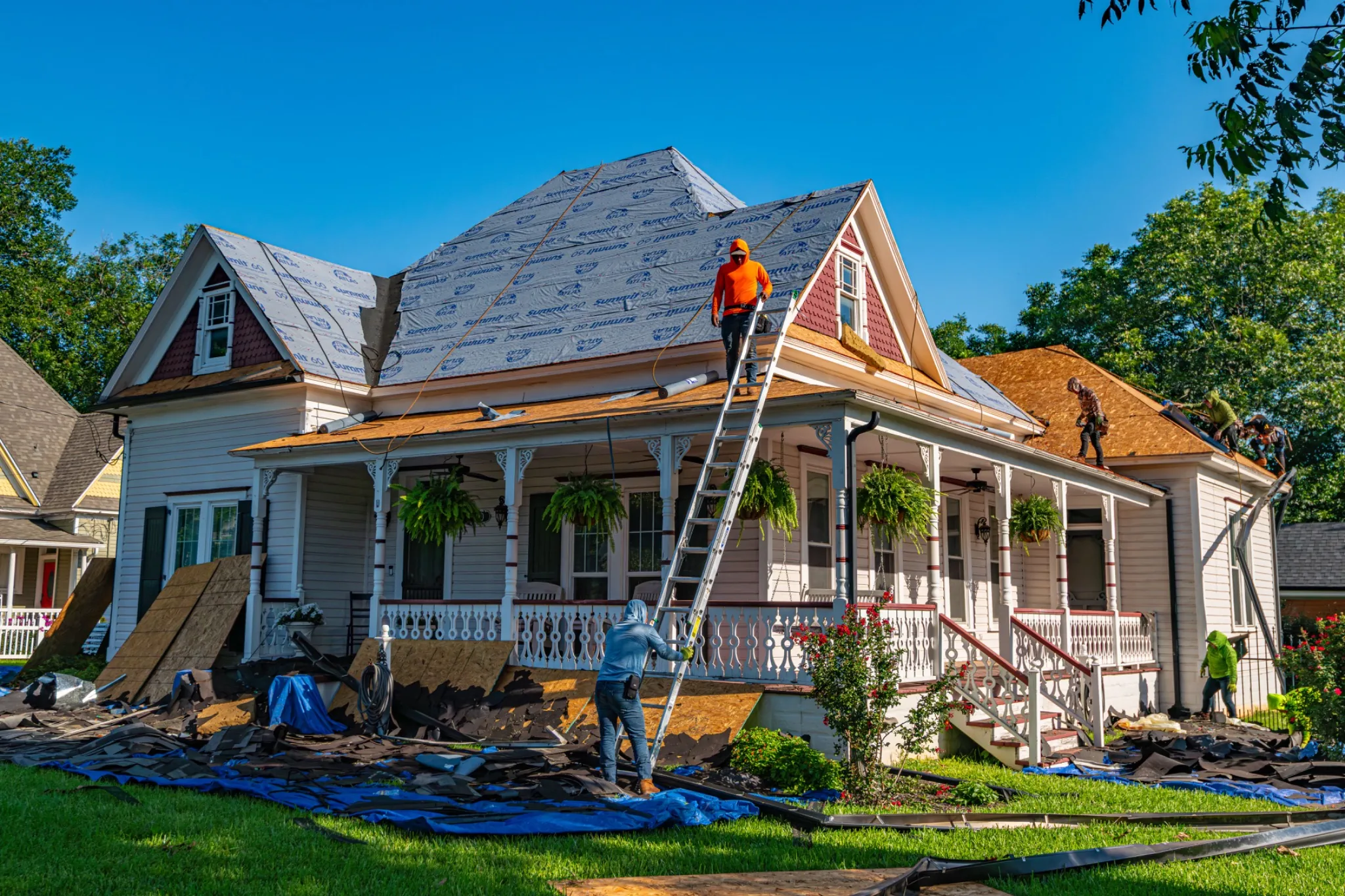 This one's asphalt shingle underlayment ("Atlas Summit 60" is a shingle-brand synthetic underlayment) — not related to Enviroshake. I'd leave the brand out here. Roofing crew stripping and re-roofing a Victorian-style home, with workers on ladders and rooftops installing synthetic underlayment over a gabled roof, surrounded by torn-off shingles and debris on the lawn below.