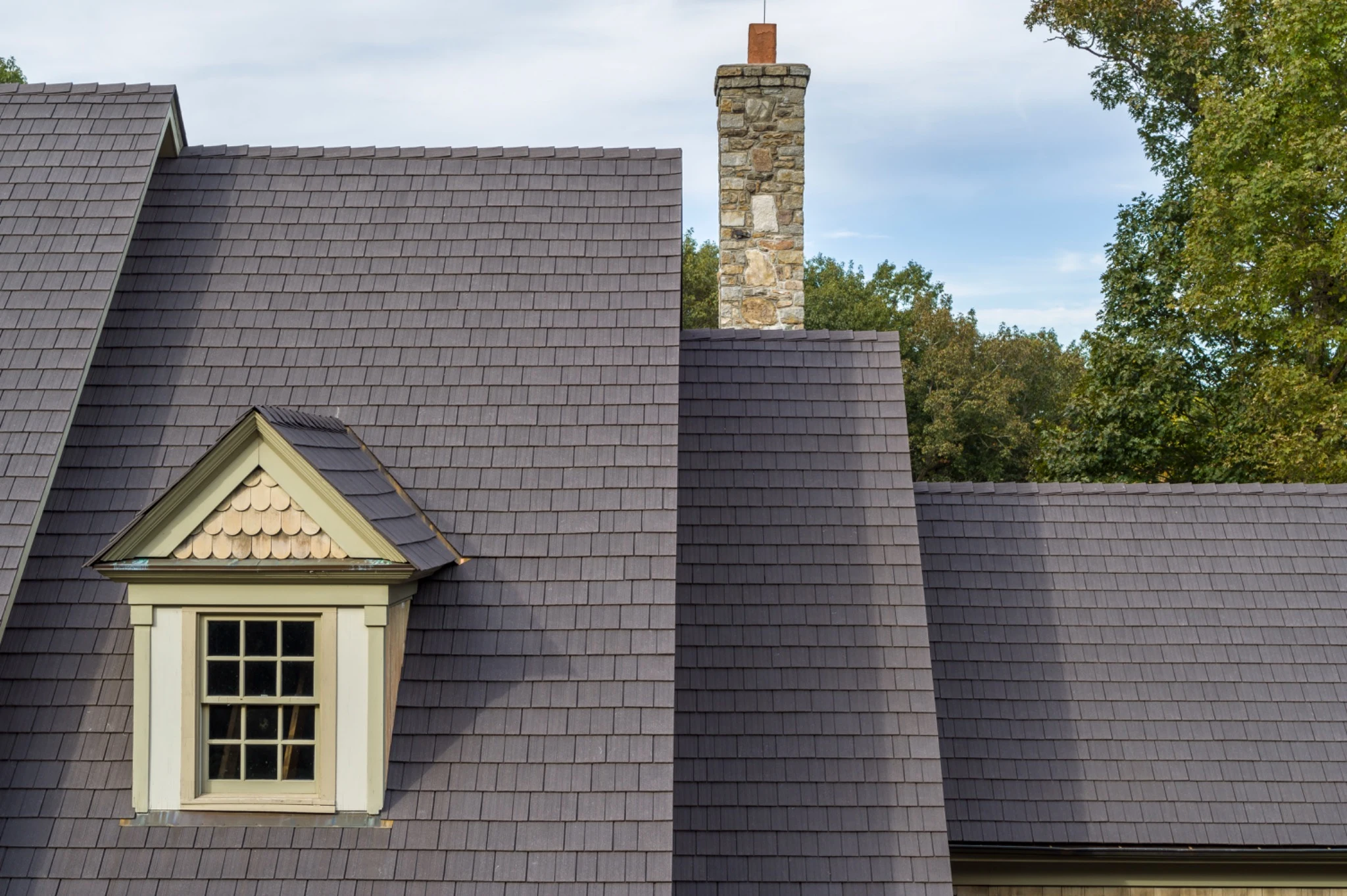 Close-up of a gabled roof in aged cedar Enviroshake composite shake, with a dormer window and copper-scaled gable trim, next to a stone chimney and tree line.