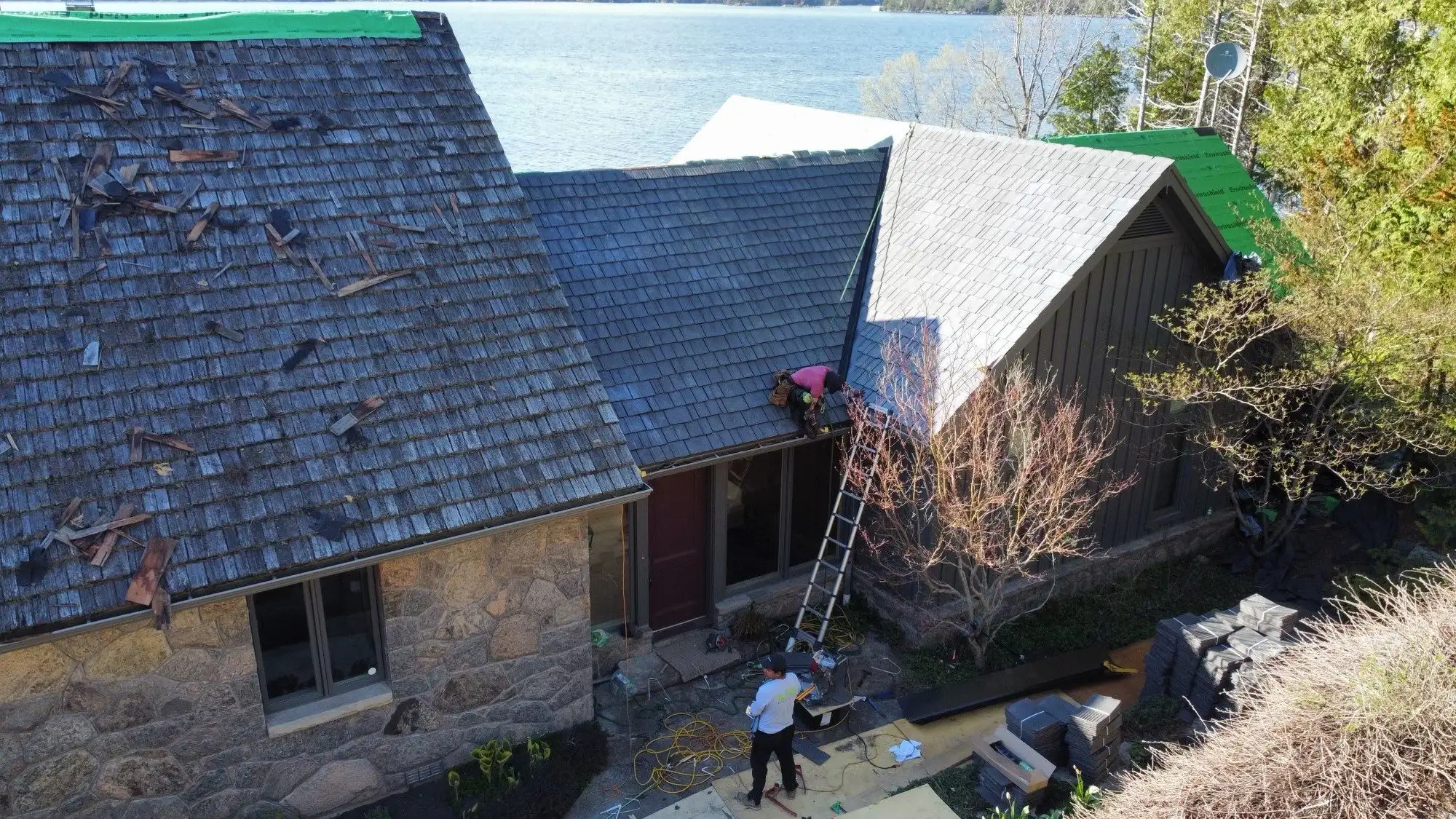 Aerial view of a stone lakeside home mid-reroofing, with weathered gray cedar shakes partially removed and stacks of new Enviroshake composite shingles staged for installation, green Enviroshield underlayment visible, and a worker on a ladder, overlooking a lake.