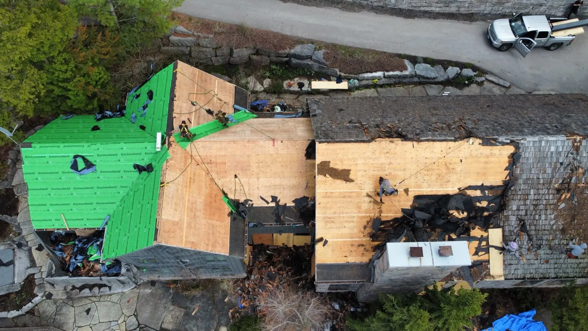 Aerial view of a stone lakeside home mid-reroofing, with weathered gray cedar shakes partially removed and stacks of new Enviroshake composite shingles staged for installation, green Enviroshield underlayment visible, and a worker on a ladder, overlooking a lake.