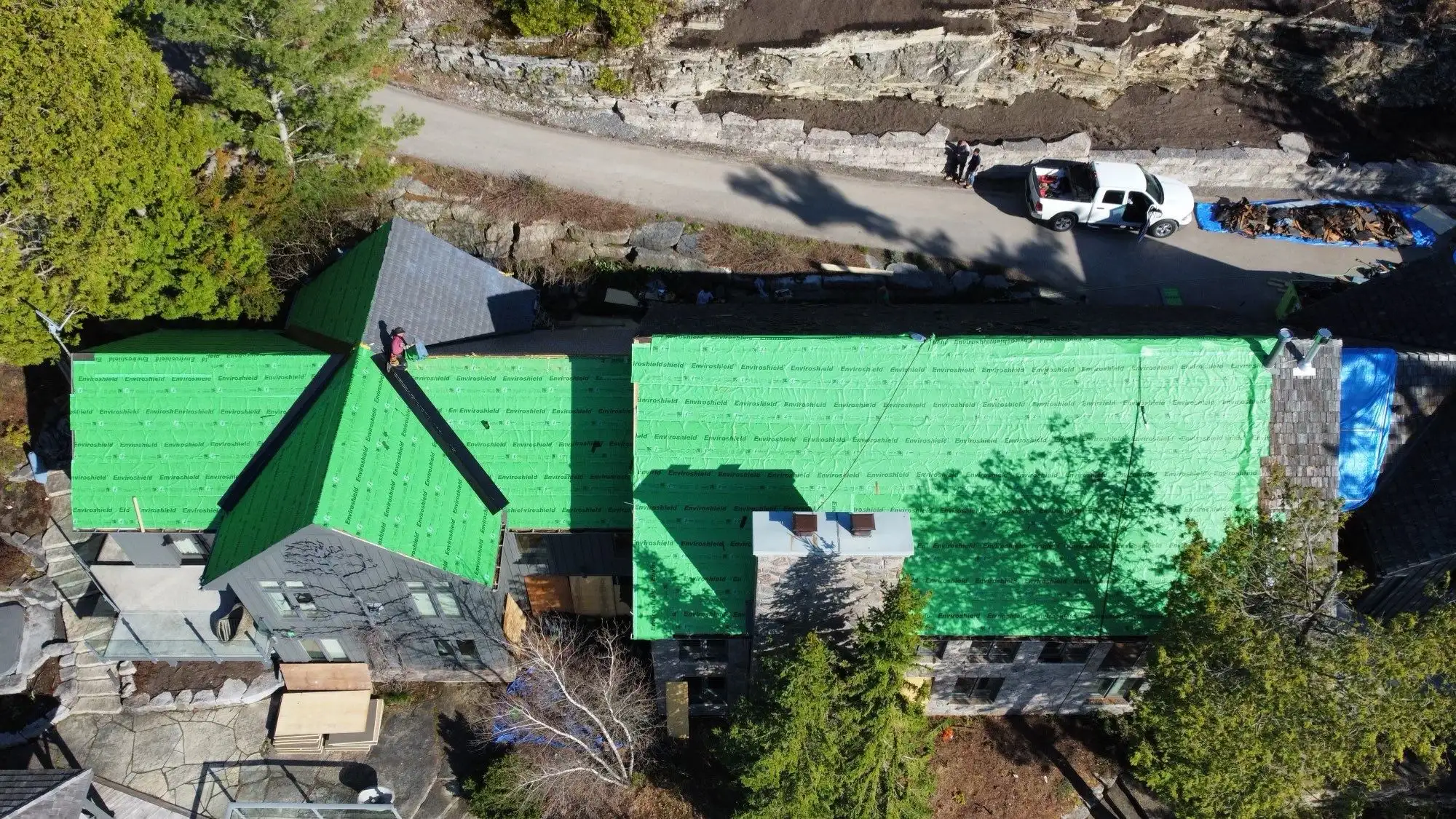 Aerial view of a stone lakeside home mid-reroofing, with weathered gray cedar shakes partially removed and stacks of new Enviroshake composite shingles staged for installation, green Enviroshield underlayment visible, and a worker on a ladder, overlooking a lake.