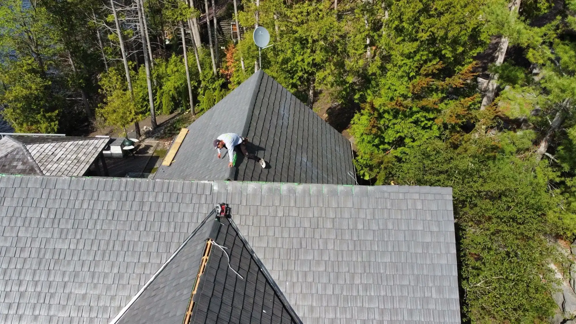 Aerial view of a worker installing a gray Enviroshake composite roof on a multi-hip roofline, surrounded by dense forest near a lake.