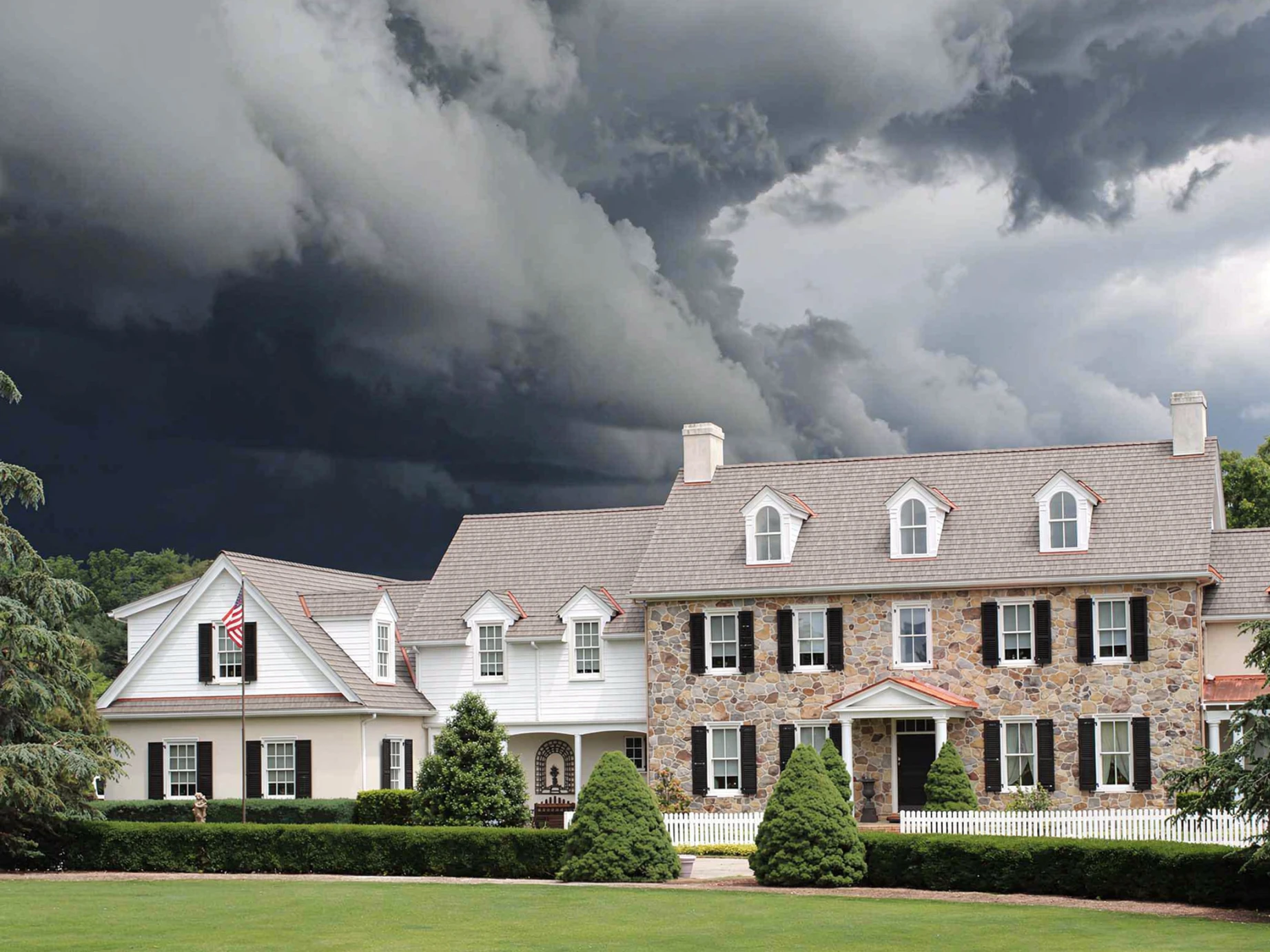 Large stone and white colonial home with an aged cedar Enviroshake composite roof, arched dormer windows, and a white picket fence, set beneath a dramatic dark storm sky.