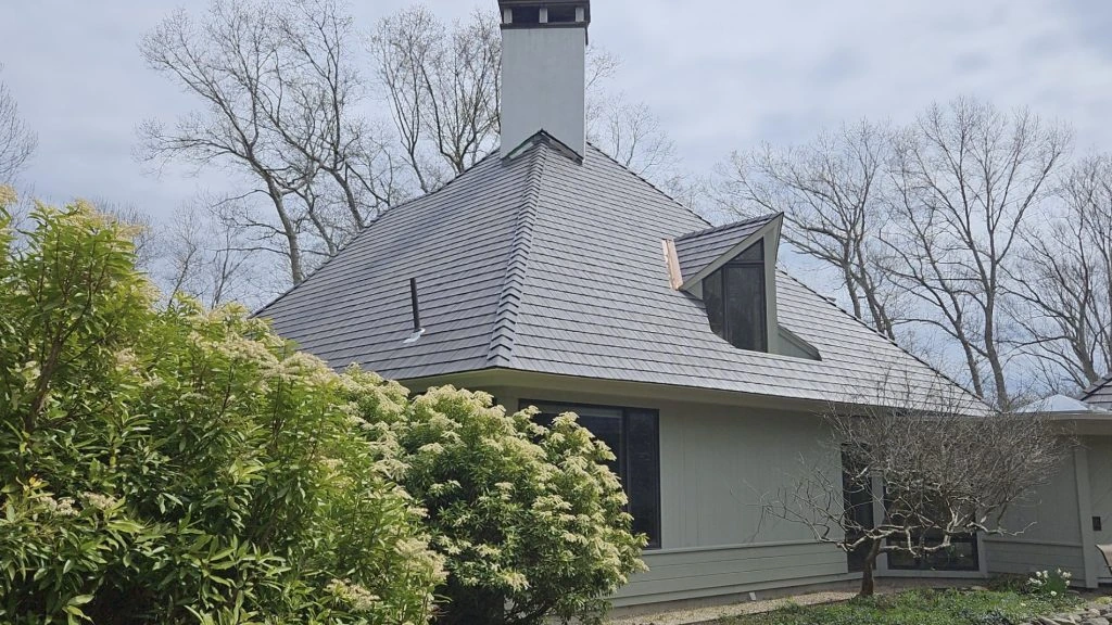 Modern gray shingle-style home with a steeply pitched Enviroshake composite roof, tall floor-to-ceiling windows, a chimney, and a rooftop deck, set among leafless trees with landscaped stonework.
