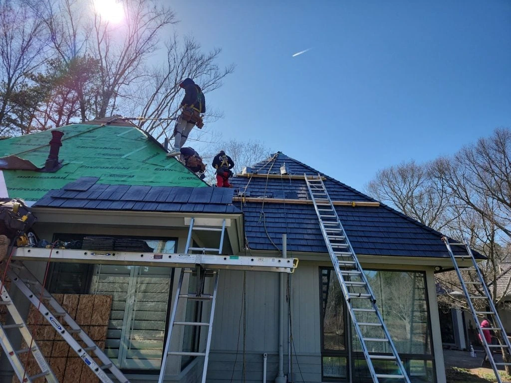 Roofing crew installing a dark blue-gray Enviroshake composite roof on a steeply pitched hipped roof, with green Enviroshield underlayment visible and multiple ladders set against the home, framed by bare trees.