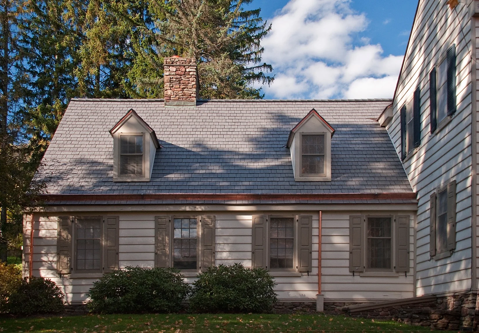 Single-story white colonial addition with a gray Enviroshake composite roof, two gabled dormers, a stone chimney, and shuttered windows, set beside a larger white farmhouse.