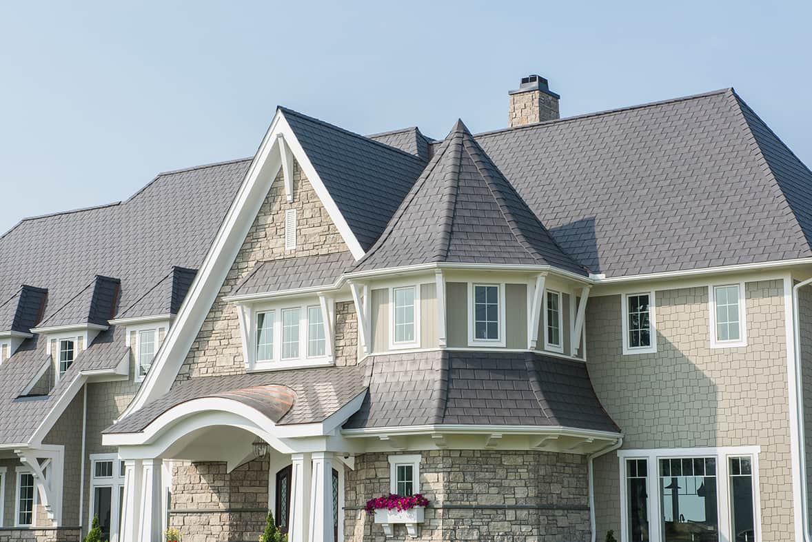 Large stone and shingle-style home with a gray Enviroshake composite roof, a round turret with a conical roofline, gabled dormers, and a curved copper entrance canopy.