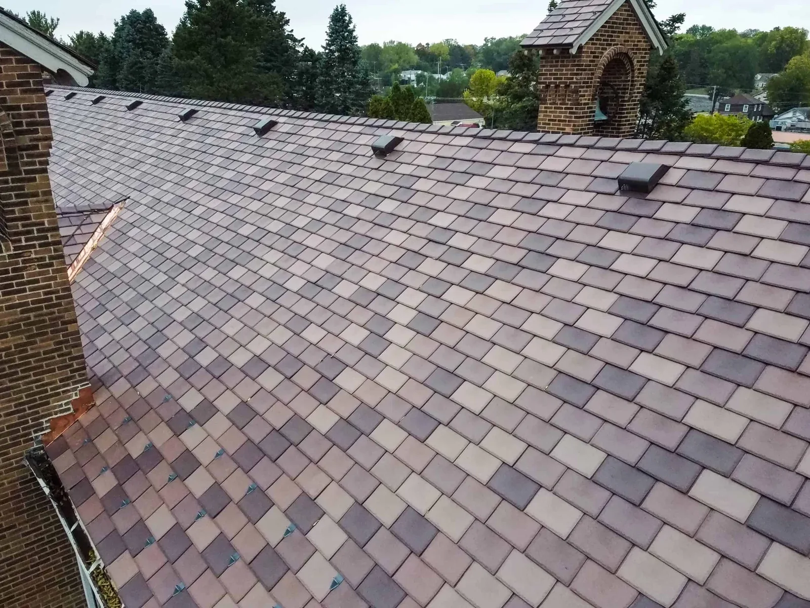 Aerial close-up of a multitone Enviroslate composite slate roof in blended mauve and gray tones on a brick church building, with roof vents and a bell tower, overlooking a wooded neighborhood.