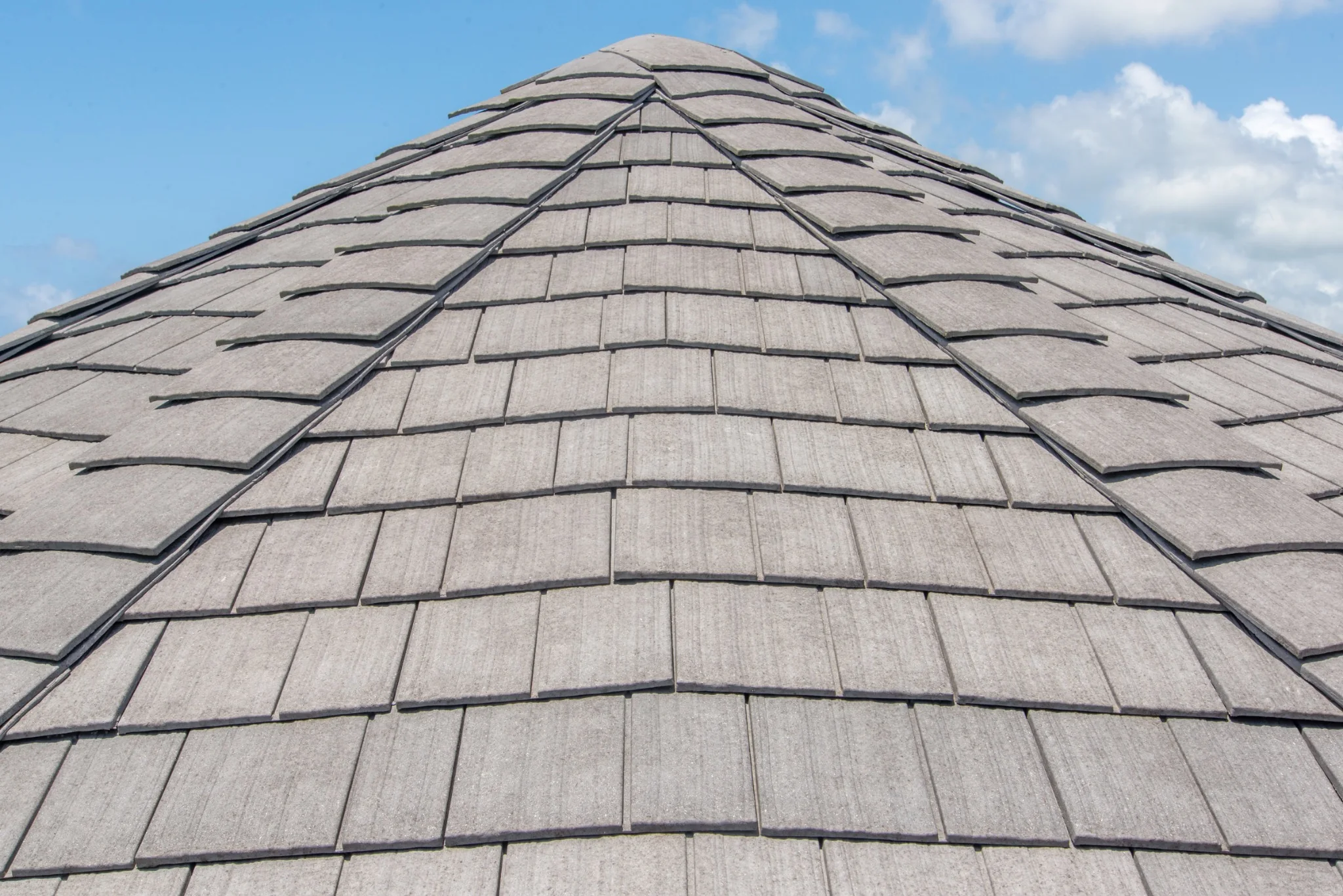 Close-up of a gray Enviroshake composite roof rising to a pyramidal peak, showing consistent shingle texture and staggered rows against a partly cloudy sky.