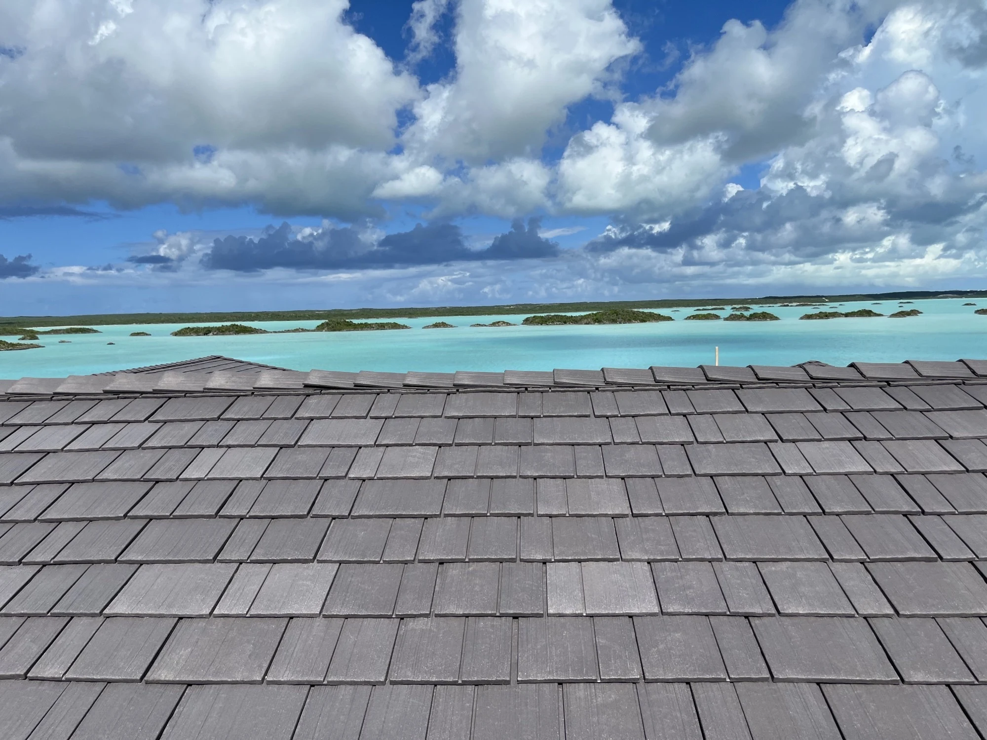 Close-up of a gray Enviroshake composite roof with a ridge cap, overlooking a turquoise lagoon dotted with small green islands under a partly cloudy sky.