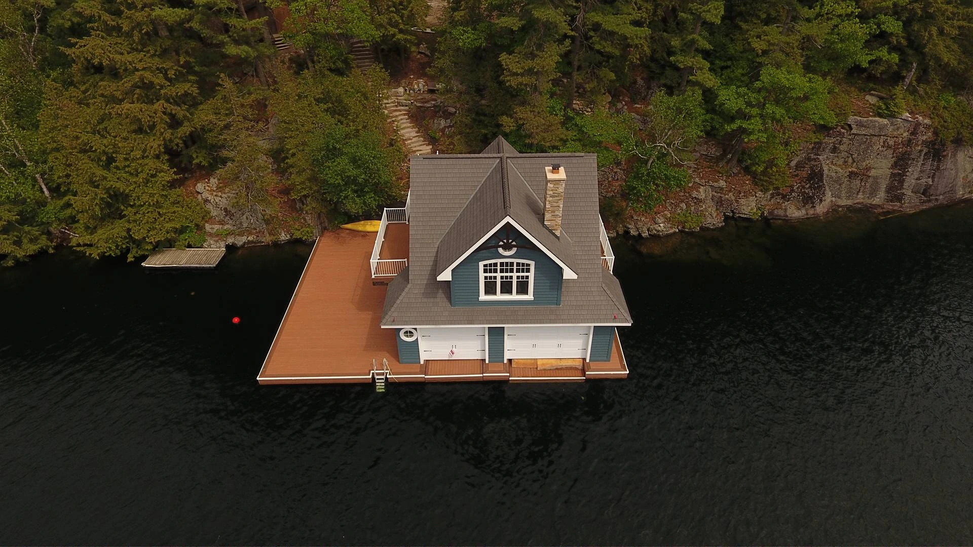 Aerial view of a blue boathouse with an aged cedar Enviroshake composite roof and white garage doors, built on a dock at the edge of a dark lake, surrounded by pine trees and rocky shoreline.