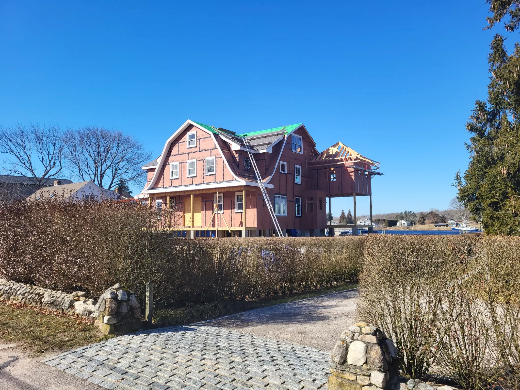 This one's under construction with just sheathing and underlayment exposed — no Enviroshake visible yet. I'd hold off on the brand mention. Home under construction with a gambrel roof, exposed plywood sheathing, and green underlayment, framed by a hedge-lined stone entrance and bare trees.