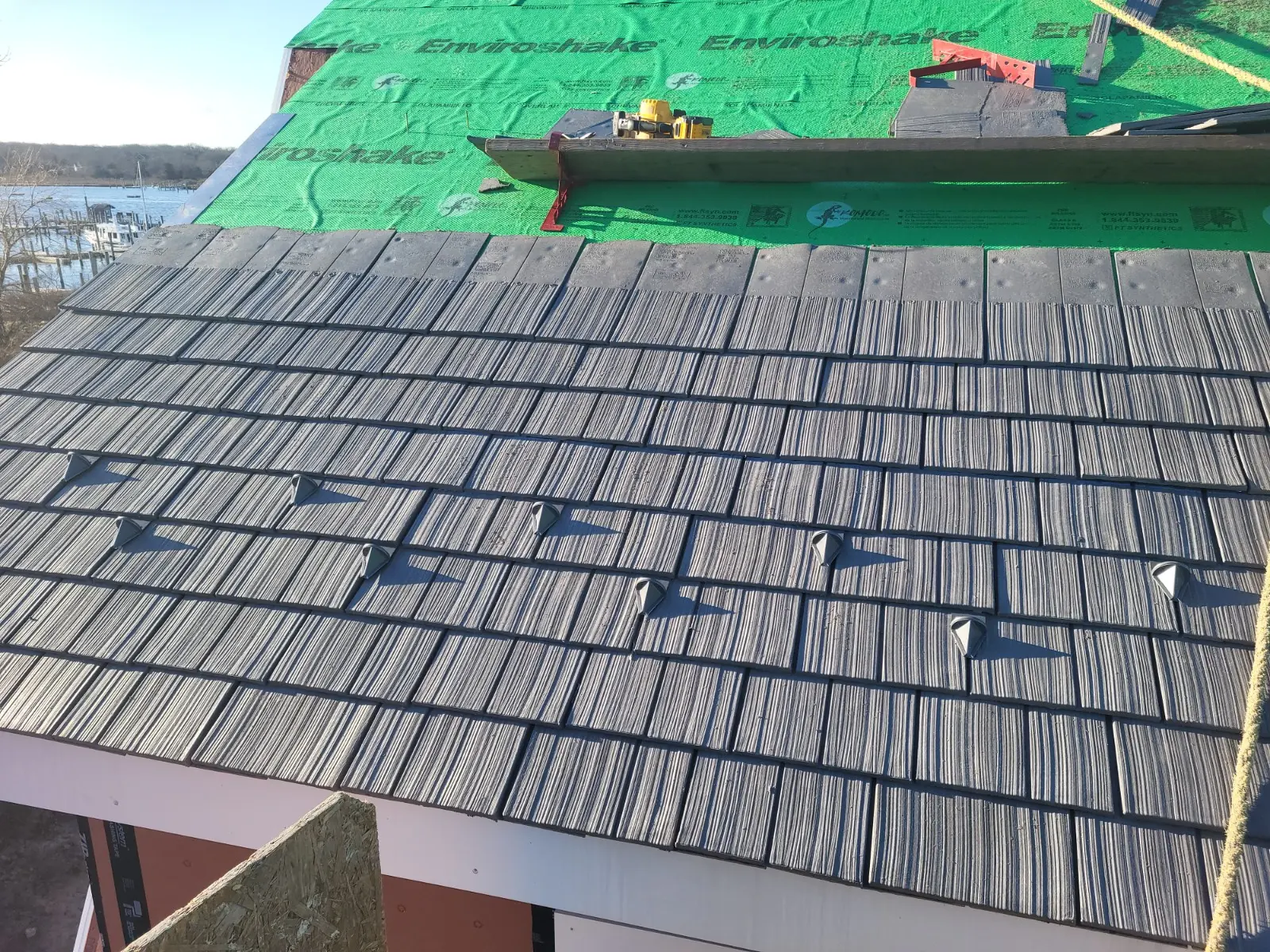 Close-up of a gray Enviroshake composite roof mid-installation, with snow guards, exposed green Enviroshield underlayment, and roofing tools, overlooking a waterfront marina.