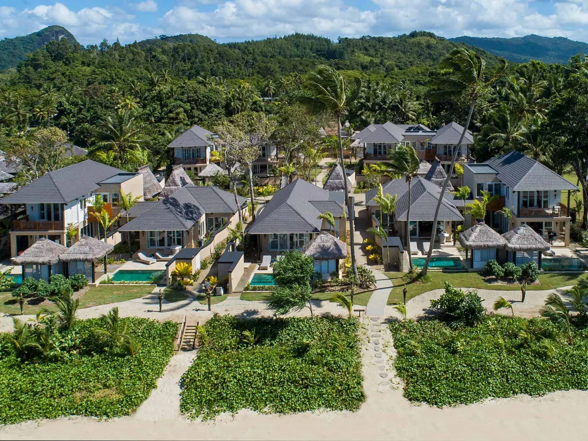 Aerial view of a tropical beachfront resort featuring Enviroshake Aged Cedar composite roofing across multiple villas, surrounded by lush palms, private pools and dense island vegetation.