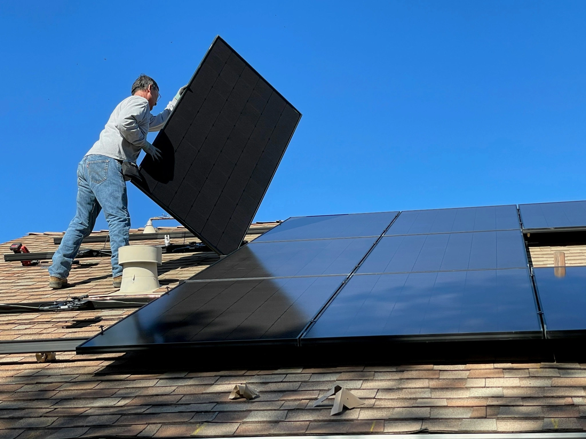 Installer mounting solar panels on an asphalt shingle roof under a clear blue sky.