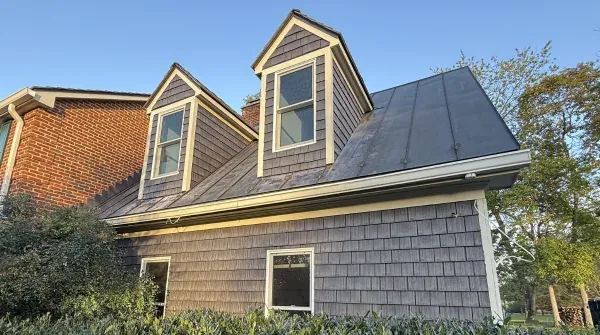 Side view of a house with weathered wood shake siding and roofing, dormer windows, and solar panels installed on the main roof slope