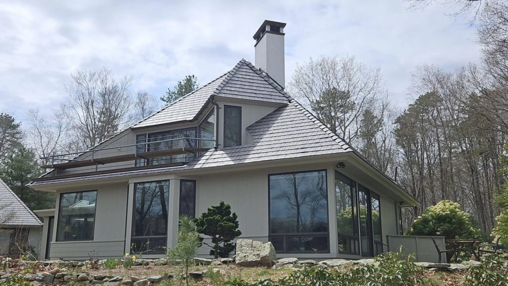 Modern gray shingle-style home with a steeply pitched Enviroshake composite roof, tall floor-to-ceiling windows, a chimney, and a rooftop deck, set among leafless trees with landscaped stonework.