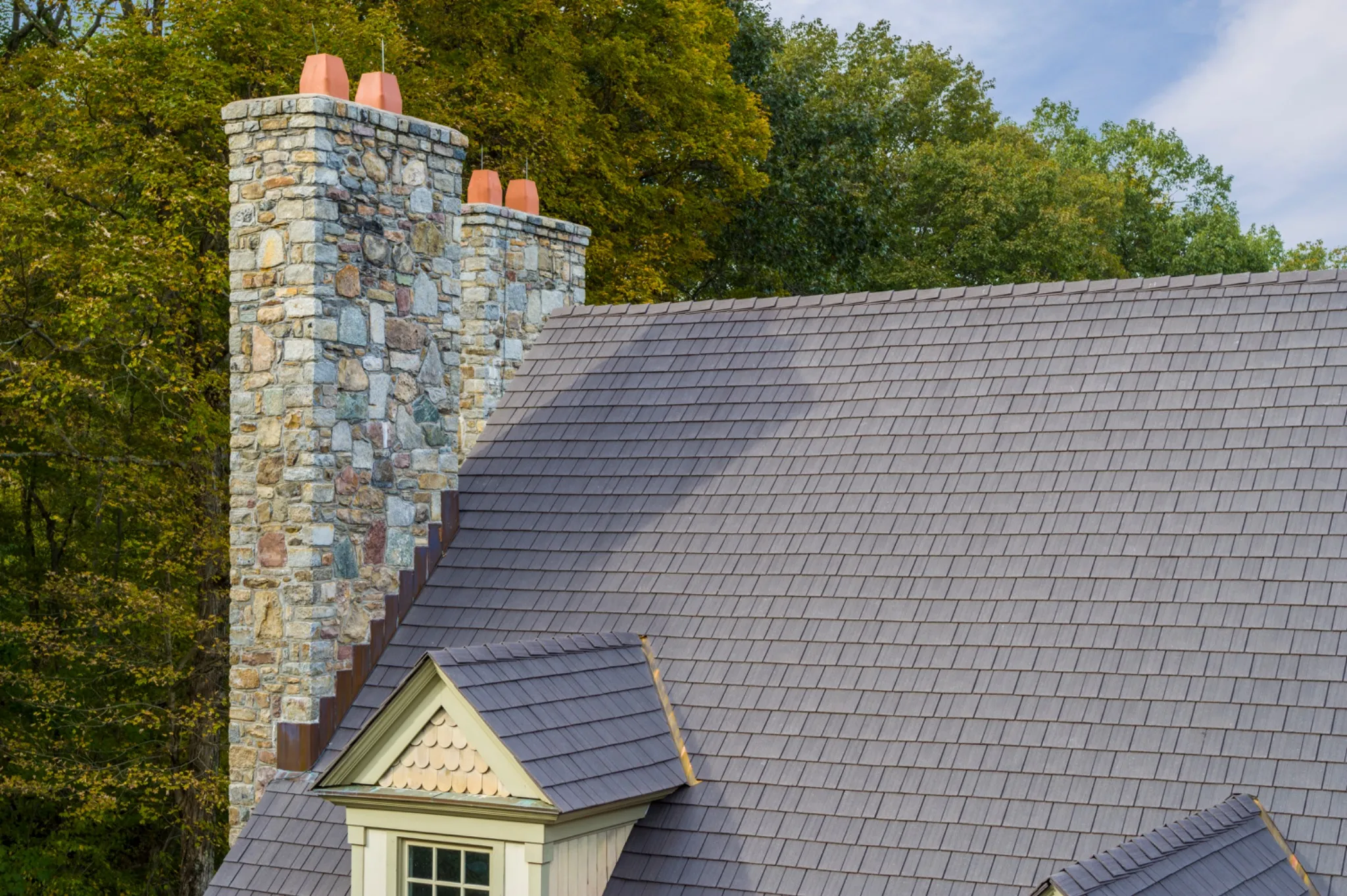 Close-up of an aged cedar Enviroshake composite roof with a small dormer, next to twin stone chimneys with terra cotta chimney pots, set against a backdrop of fall foliage.