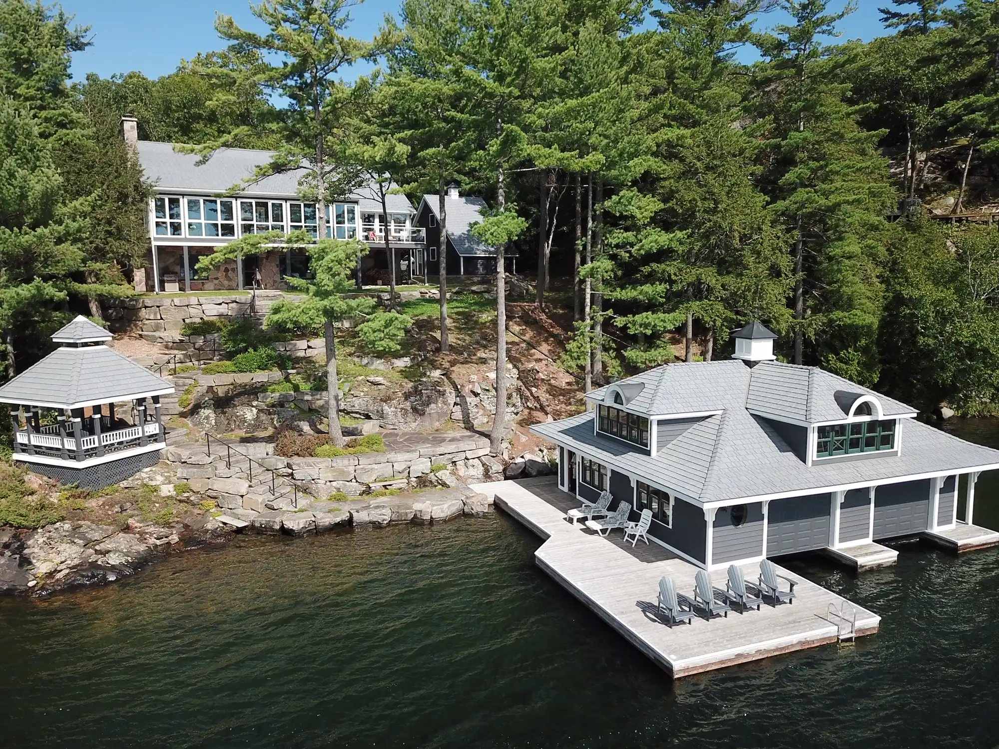 Aerial view of a lakeside property with a silvered cedar Enviroshake composite roof on the main house, gazebo, and boathouse, connected by a rocky shoreline and dock with Adirondack chairs, surrounded by dense pine trees.