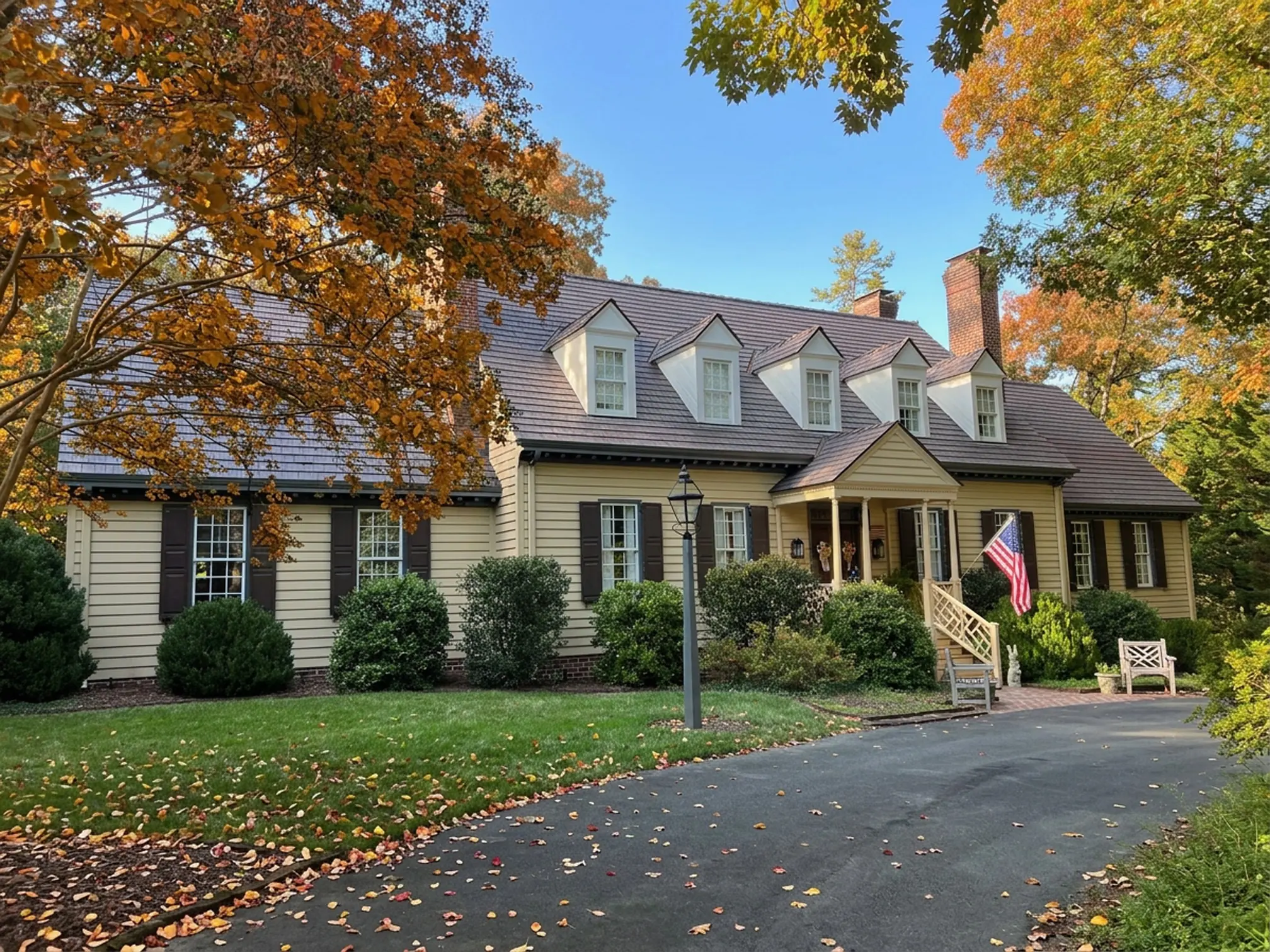 Colonial-style yellow home with an aged cedar Enviroshake composite roof, five dormer windows, brick chimneys, and an American flag by the front porch, framed by fall foliage.